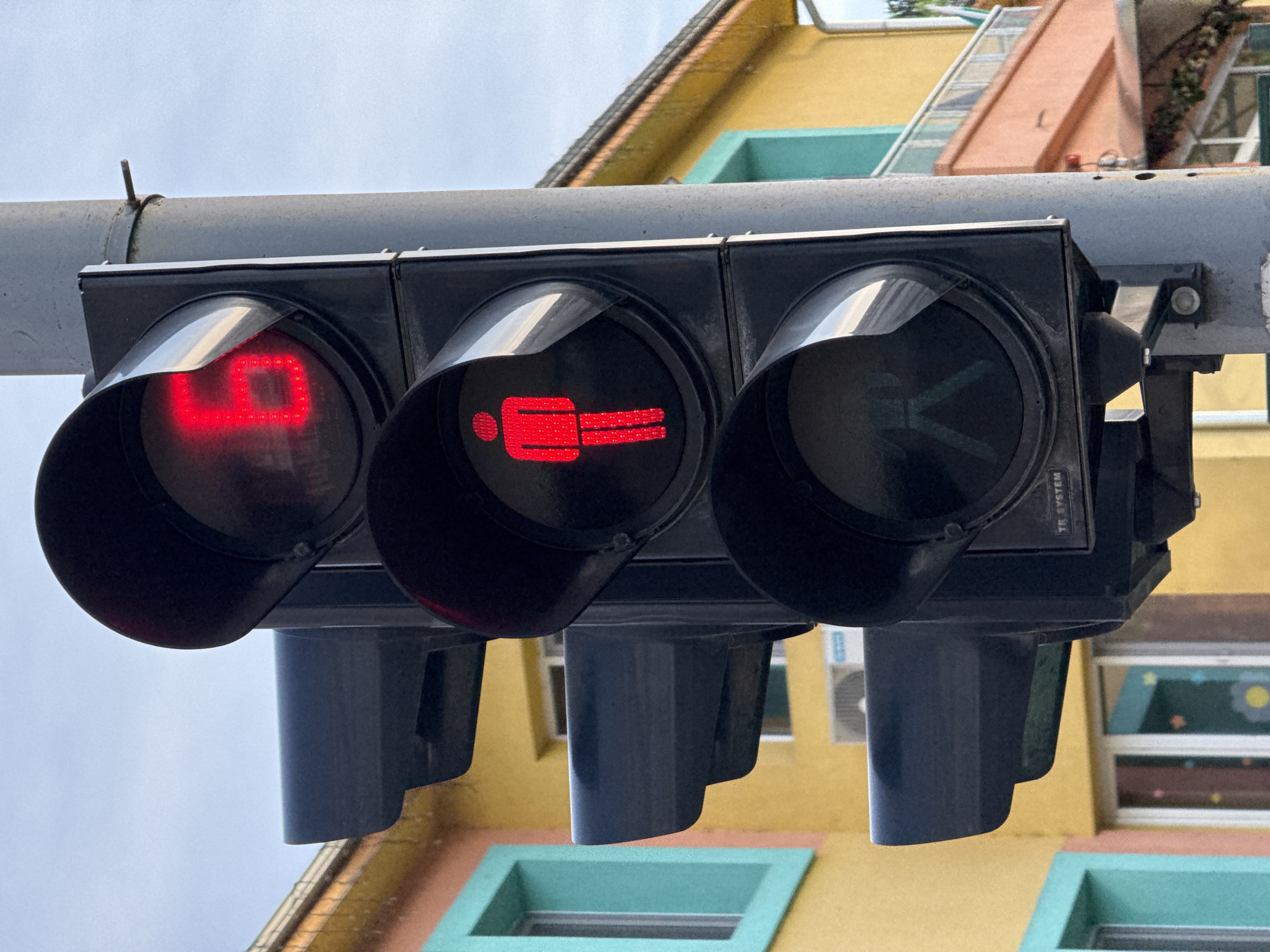 The image shows a traffic light for pedestrians in Lovech, Bulgaria. The signal on the left displays the number "9" in red, indicating the remaining time to cross. The middle signal shows a stationary red pedestrian figure, signaling "Do Not Walk." The right signal is dark. The traffic light fixture is black, and mounted on a grey pole. The background reveals a multi-story building with yellow walls and green window frames.