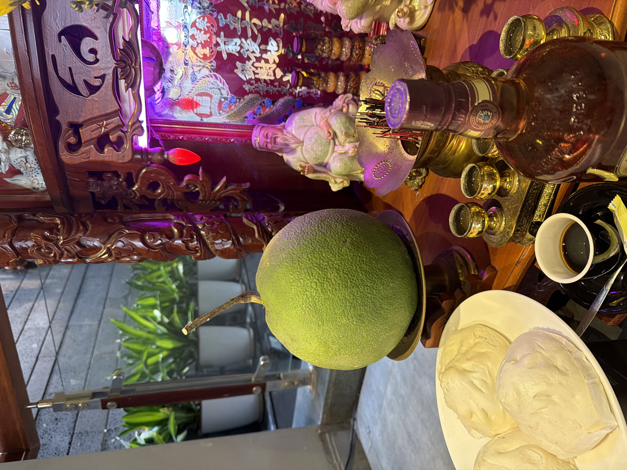 In a room in South Quy Nhon Ward, Vietnam, a religious altar is set up. A large, green pomelo rests on a brass stand to the left of the frame. In front of the altar, a white plate holds three steamed buns. To the right of the plate, a small white cup filled with dark liquid, likely coffee, sits on a black saucer, accompanied by a spoon. A bottle of amber liquid, possibly whiskey, stands prominently in the foreground, its label partially visible. The altar itself features an ornate wooden frame, illuminated by purple and red lights. Within the altar, a seated figurine of a man with a long white beard is visible, along with a brass incense burner filled with incense sticks. Various decorative items, including bottles and a red banner with Chinese characters, are also present on the altar. In the background, through a glass pane, several potted plants with vibrant green leaves can be seen. The scene appears to be indoors, with tiled flooring visible in the foreground.