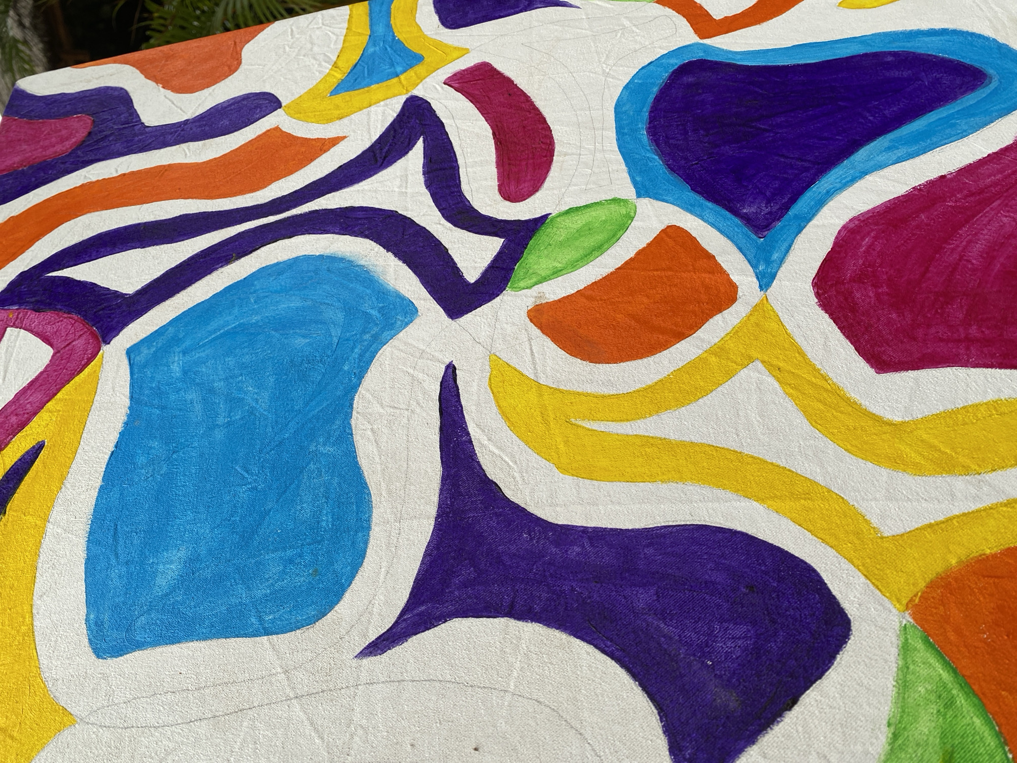 The image displays a close-up, angled view of a colorful abstract painting on a white canvas. The painting features bold, organic shapes in various bright colors, including blue, purple, yellow, orange, green, and pink. The brushstrokes are visible, giving the artwork a textured appearance. The background of the painting is white, with some visible canvas weave and faint pencil lines suggesting a sketch underneath. The painting appears to be outdoors, with a hint of green foliage visible in the upper right corner, suggesting it might be situated in a garden or natural setting. There is no information within the image that would allow for its location to be identified in Gura Top, Nigeria.