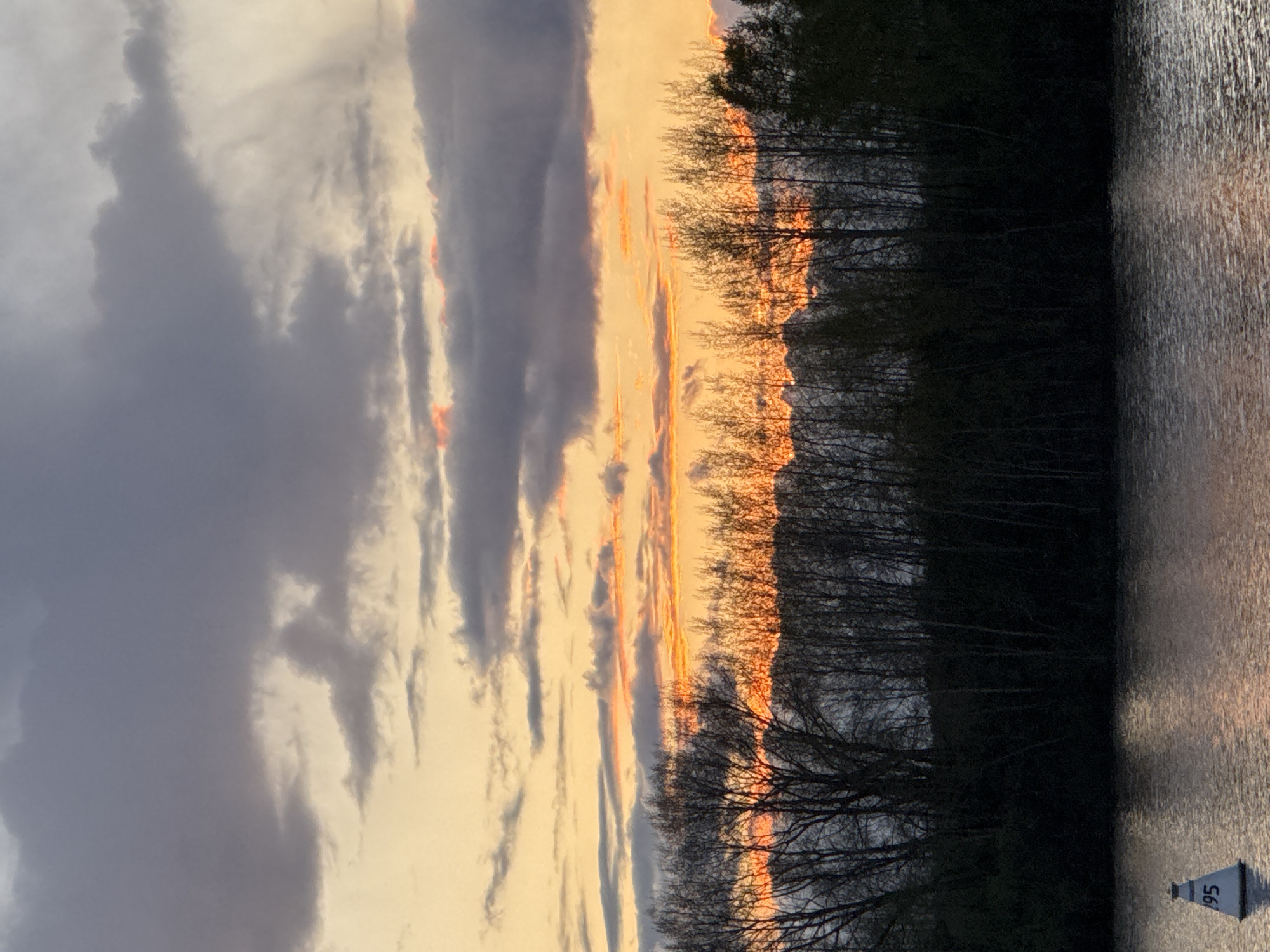 The image shows a sunset scene in Chaikovsky, Russia. A body of water, possibly the Kama River, reflects the sky. A line of trees silhouettes against the setting sun. The sky is filled with clouds, some dark and heavy, others reflecting the orange and yellow light of the setting sun. In the bottom right corner, a white channel marker with the number "95" is visible in the water. The image is oriented vertically.