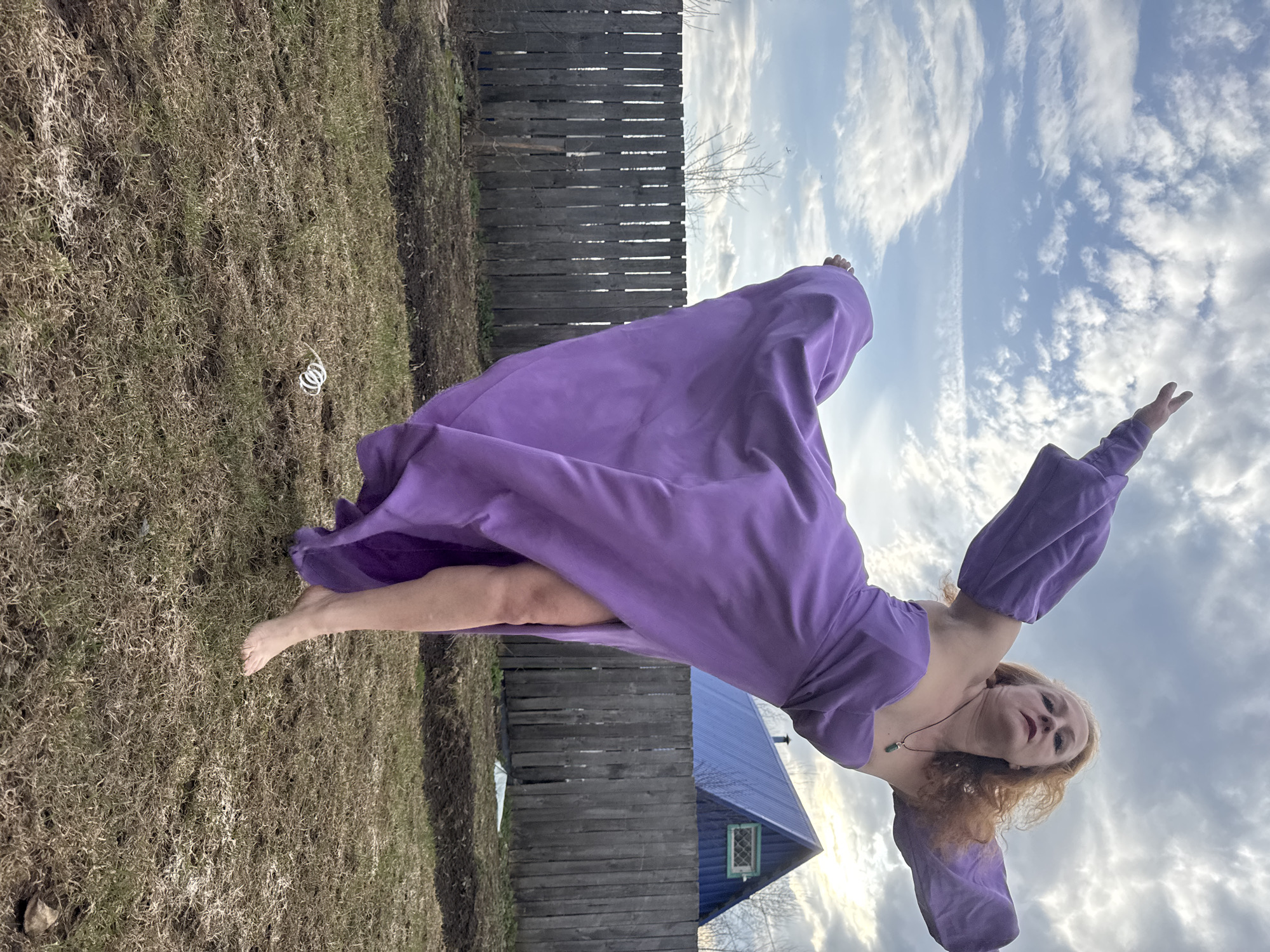 An outdoor, rotated full-shot captures a woman in mid-air, performing a graceful leap or dance against a partly cloudy sky in Chaikovsky, Russia. The woman, with reddish-blonde curly hair, wears a flowing, long purple dress with puffy sleeves and an off-the-shoulder neckline. She is barefoot, with one leg bent and the other extended, and her arms raised outwards, embodying a sense of freedom and movement. A necklace with a small green pendant adorns her neck. Her expression is focused and serene.
The setting consists of sparse, dry green and brown grass in the foreground, where a small white coiled object is visible. Behind her, a weathered wooden fence runs horizontally across the midground. Beyond the fence, a small, rustic house with a striking bright blue corrugated roof and a light-framed window is partially visible. The sky features scattered white and grey clouds against patches of light blue, suggesting daytime, possibly late afternoon, with diffused light. No text is visible in the image.