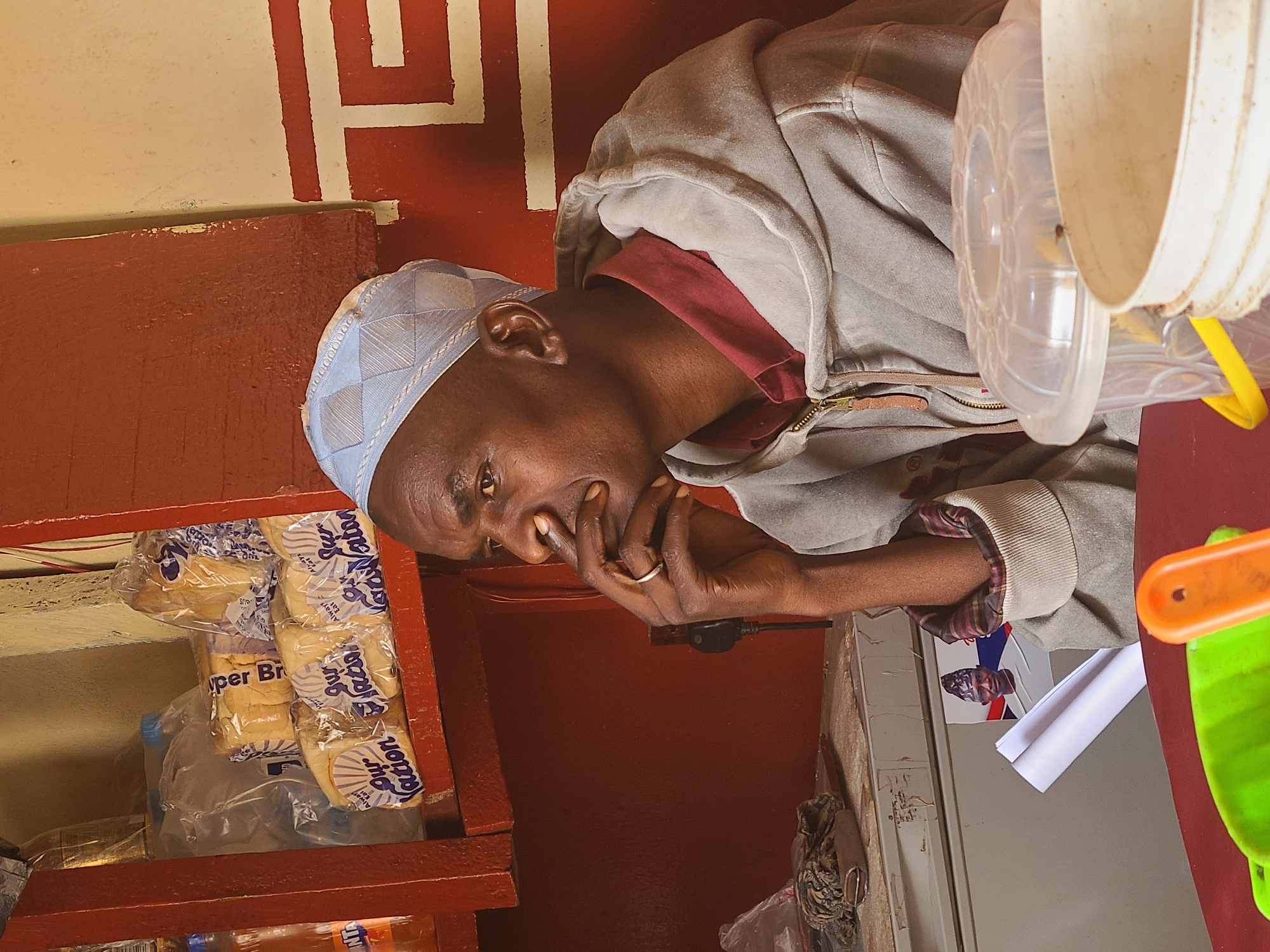 A medium shot depicts a dark-skinned man wearing a light blue kufi and a gray hooded sweatshirt over a maroon collar, positioned slightly to the right of the frame, as though he is sitting on a shelf in a market stall. His right hand is brought up to his face and he appears to be in thought. The man is located on a shelf and in front of him are several loaves of bread. The setting seems to be a small shop with red walls and painted patterns, a plastic bucket and a poster featuring another person's face. The location in the city is Unknown, Unknown.