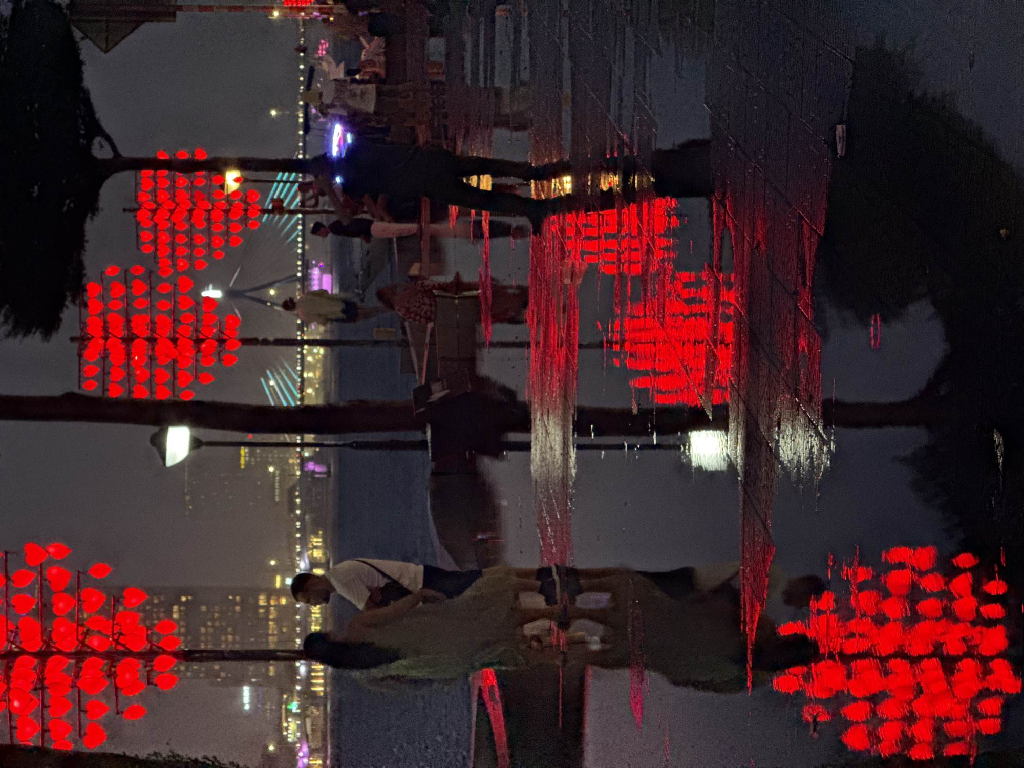 At night, the city of Da Nang, Vietnam, is illuminated by numerous red heart-shaped lights strung on trees, reflecting vibrantly on wet pavement and in puddles. The scene is likely along a waterfront promenade or a pedestrian area, given the presence of water and the distant city lights and bridge. In the foreground, a couple stands close together, their silhouettes and reflections visible in the wet ground. Other figures, some blurred, are also present in the background, suggesting a public space. The ambiance is romantic and slightly mysterious due to the darkness and the glowing lights. The image captures a moment of urban beauty in the rain.