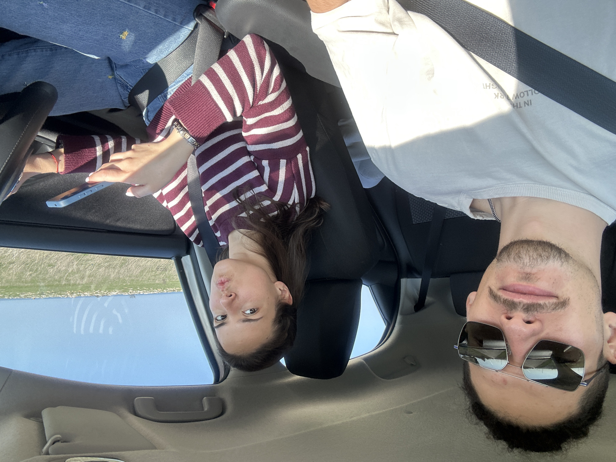 Two people are inside a car. A woman with long, dark hair is in the passenger seat, wearing a striped maroon and white sweater, and holding a smartphone. A man is in the driver's seat, wearing sunglasses and a light-colored t-shirt. The car is near a coastline of Kamen bryag, Bulgaria, visible through the window.
