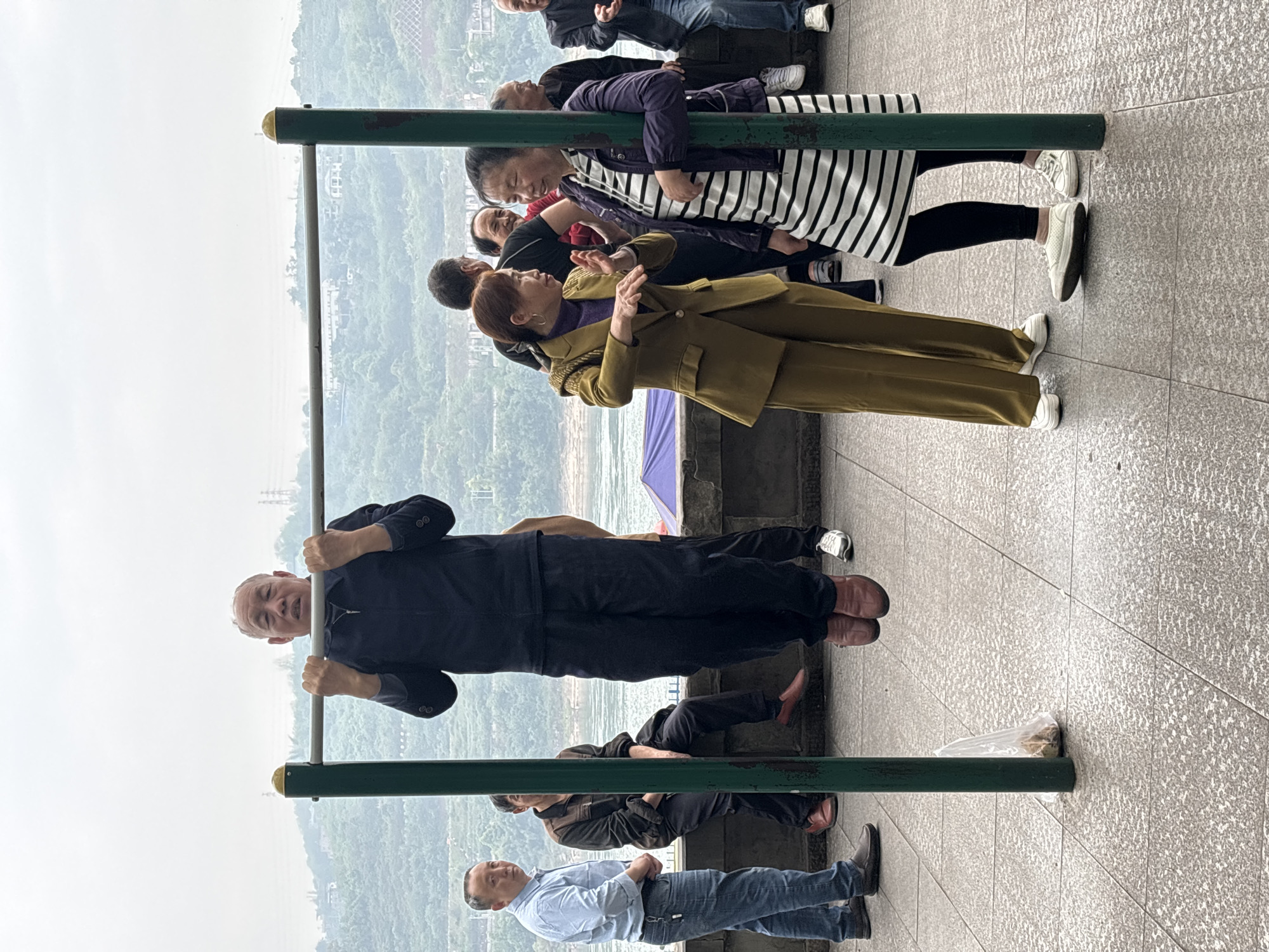 A man in a dark blue suit and reddish brown shoes is performing a pull-up on a green metal bar in what appears to be an outdoor public space in Jiangjin, China. Several people are standing nearby, observing and seemingly conversing. One woman in a mustard-colored suit and white shoes is gesturing with her hands while talking to another woman in a striped shirt. Other individuals are visible in the background, including a man in a light blue shirt and jeans standing at the base of the metal bar. The surroundings include a view of a river and hilly landscape.