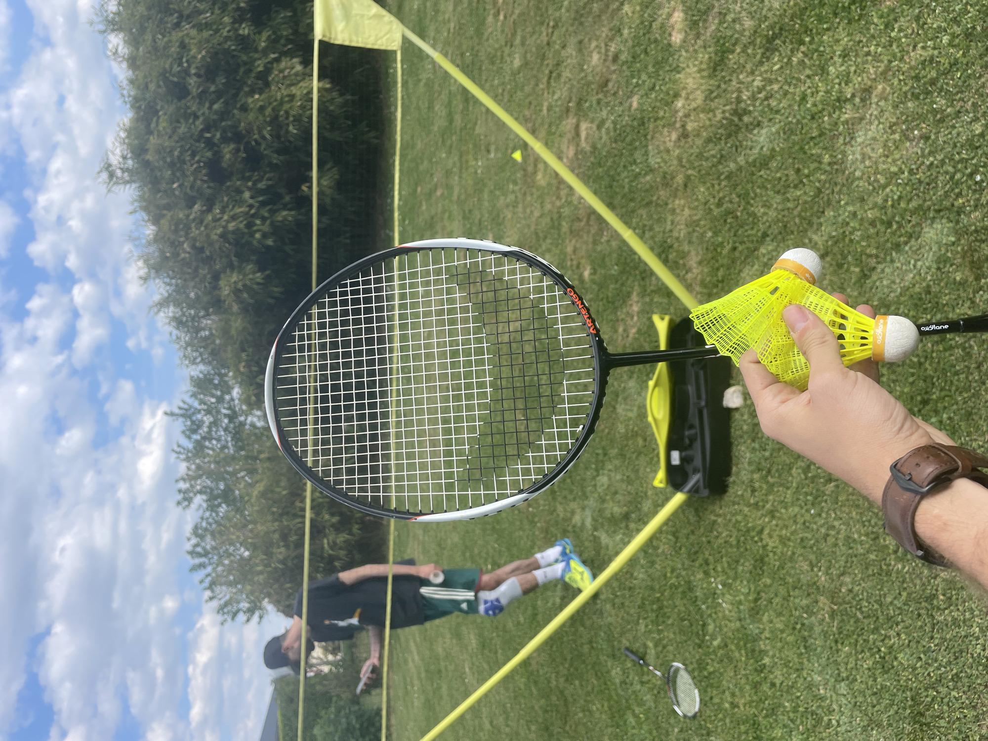 The image captures an outdoor badminton game or practice session on a sunny day in La Chapelle-Bertrand, France. In the immediate foreground, a hand wearing a brown leather wristband holds a black and white Artengo badminton racket. The hand also grips two bright yellow shuttlecocks; one clearly shows "oxe l'ane" on its white base, and the other has "Artengo" printed on its yellow skirt.
The scene is set on a well-maintained grassy area, marked out with yellow lines and featuring a portable yellow badminton net. Across the net, a second person stands on the court, dressed in a dark t-shirt, dark green shorts with white stripes, and vibrant blue and yellow athletic shoes. This individual appears to be looking down or adjusting something, suggesting a moment between plays. Another badminton racket is visible lying on the grass in the mid-ground.
The background is dominated by a line of verdant trees under a clear blue sky with fluffy white clouds, indicating pleasant daytime weather for outdoor activities. The overall impression is one of recreational sports.
