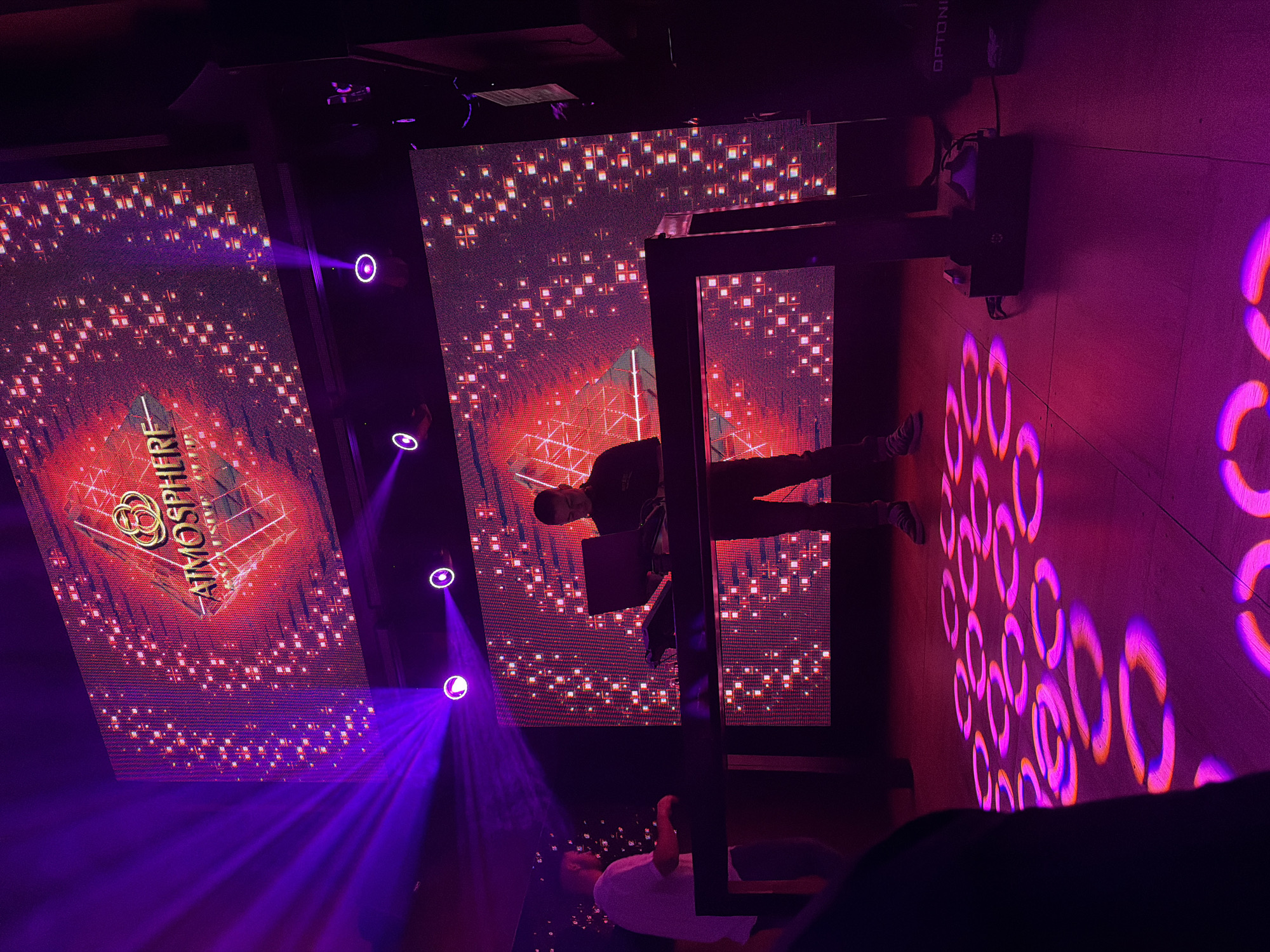 The image captures a scene inside the Atmosphere nightclub in Unknown, Unknown. A DJ is positioned behind a black structure, with a laptop in front of him. Behind the DJ, large screens display a graphic with the word "ATMOSPHERE" prominently featured. Four circular purple lights shine onto the screens, creating a dramatic effect. On the right, circular patterns of light are projected onto the wall or floor. A person in a white shirt is visible below the DJ's platform. The overall lighting is purple, contributing to the club atmosphere.