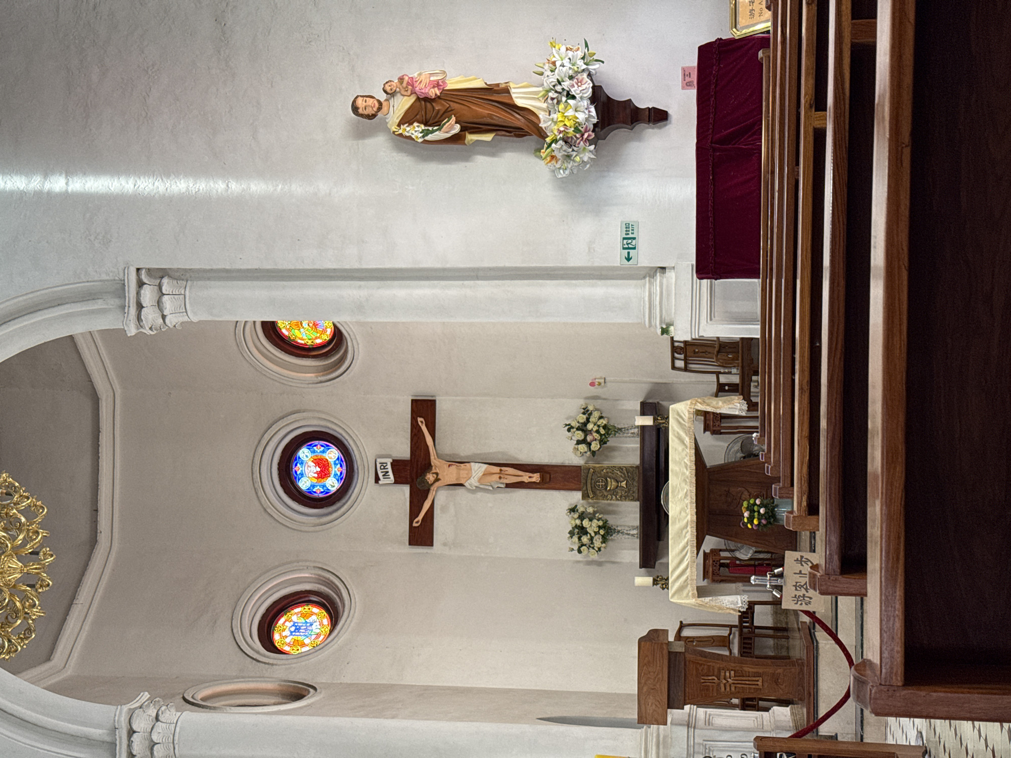 The image shows the interior of Shamian Our Lady of Lourdes Chapel in Liwan District, China. The perspective is from within the church looking toward the altar. In the foreground are rows of dark wooden pews. The nave features stained-glass windows in the upper walls and a large crucifix mounted on the wall behind the altar, flanked by floral arrangements. Above the nave on the right-side wall hangs a statue of St. Joseph holding Jesus. An exit sign is visible above the statue.