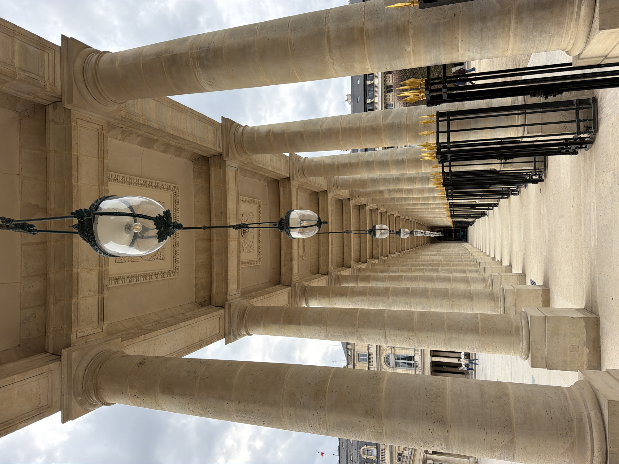 The image captures an eye-level perspective of a colonnade, likely located in Paris, France, identified by its distinctive architectural style. Rows of light beige-colored stone columns are arranged in parallel lines, creating a long, symmetrical corridor. Overhead, a series of globe-shaped lamps hang suspended by ornate metal supports. To the right, a black metal fence adorned with gold spear-shaped finials runs alongside the colonnade. In the distance, buildings with grey rooftops are visible through the spaces between the columns. The sky is overcast, casting a soft light throughout the scene. There is a couple of people on the right side in the distance.