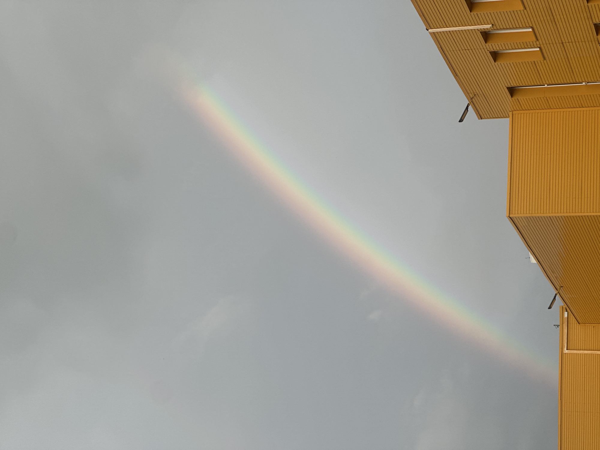 A rainbow arches across a grey, overcast sky above two sections of a modern, bright yellow building. The building's exterior features horizontal paneling and rectangular windows. The rainbow is visible as a vibrant arc of colors, with red at the top and violet at the bottom, extending from the upper left of the frame towards the lower right. No people are visible in the image. The scene appears to be an urban environment, possibly a commercial or industrial area. The location is identified as Perm, Russia.