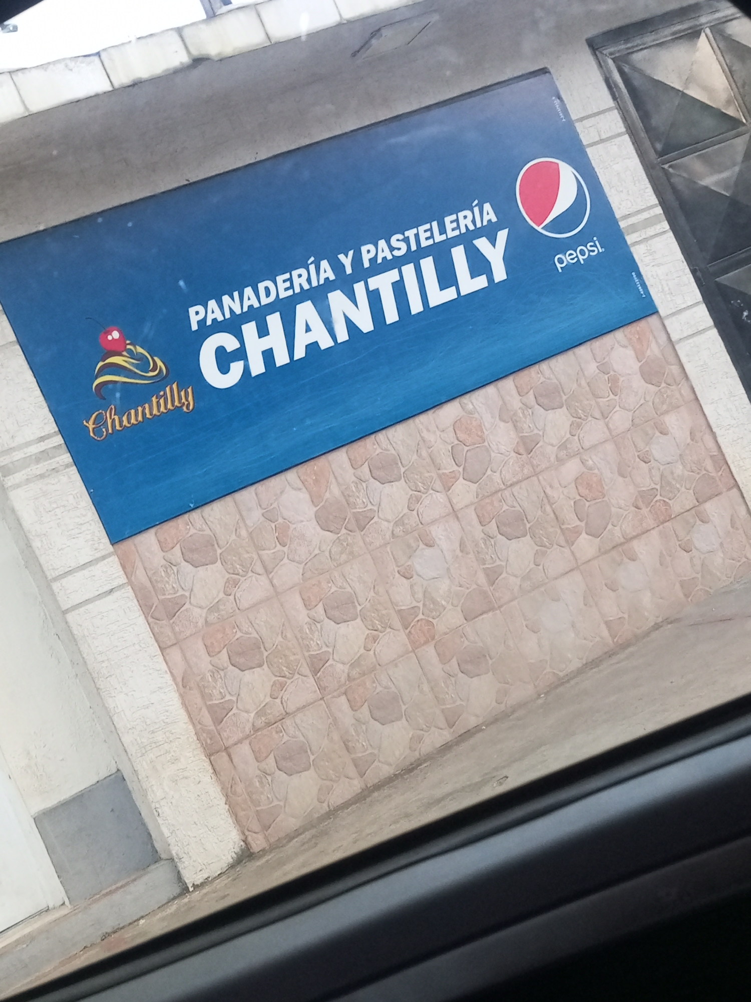 A sign for "Panaderia y Pasteleria Chantilly" with a Pepsi logo is displayed on a blue background. The sign is mounted on a building with tiled walls. To the left of the sign is a white column and a gray concrete step. To the right, a dark metal door is visible. The scene is viewed from a slightly elevated angle, suggesting it might be taken from a vehicle. The image is captured in Juan Griego, Venezuela.