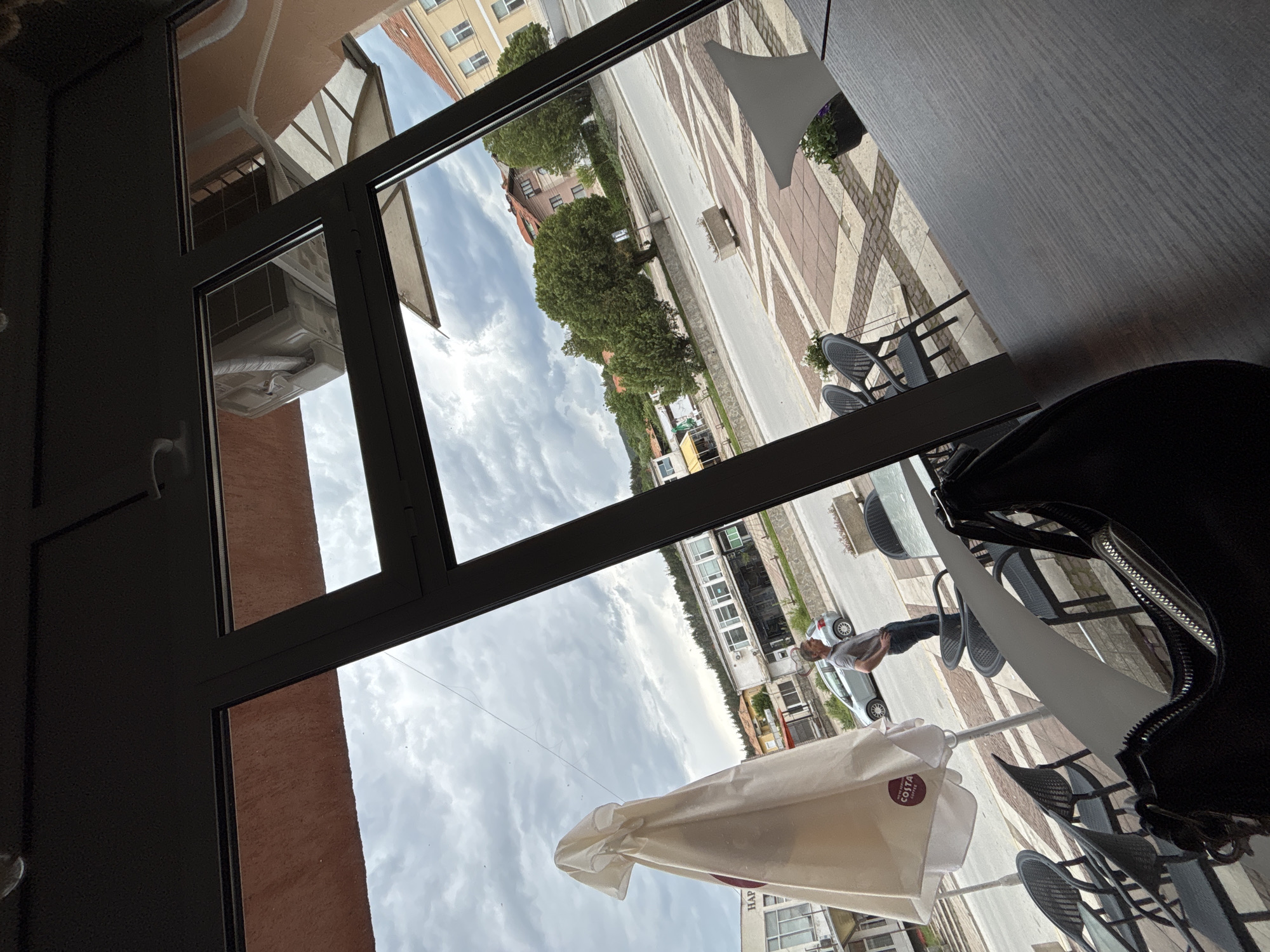 The image shows an exterior scene viewed through a window, likely in a café or restaurant in Garmen, Bulgaria. The street outside features a patterned sidewalk with decorative paving. There are trees and buildings visible on either side of the street, suggesting an urban setting. A man stands on the sidewalk near a parked car. Tables and chairs are arranged for outdoor seating, with a white umbrella partially covering the view. Inside the café, a black purse rests on a table.