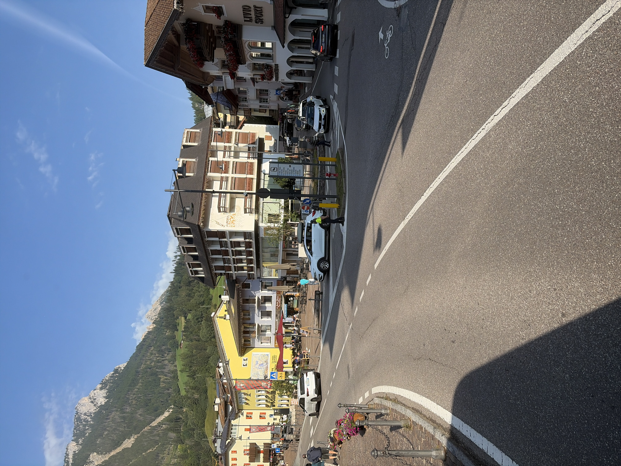 In Moena, Italy, a street intersection is depicted under a clear blue sky with a mountain range in the background. A white SUV, a dark SUV, and a white sedan are present, with the white sedan stopped at a crosswalk. A person wearing a yellow vest, likely a traffic controller or pedestrian, stands near the intersection. Several buildings line the street, displaying various architectural styles and colors, including a hotel with signs for "HOTEL AURORA" and a storefront labeled "LIVIO SPORT". Outdoor seating areas with tables and chairs are visible in front of some establishments. The road has white lane markings, including a bike lane symbol. The scene appears to be a typical sunny day in a European town.