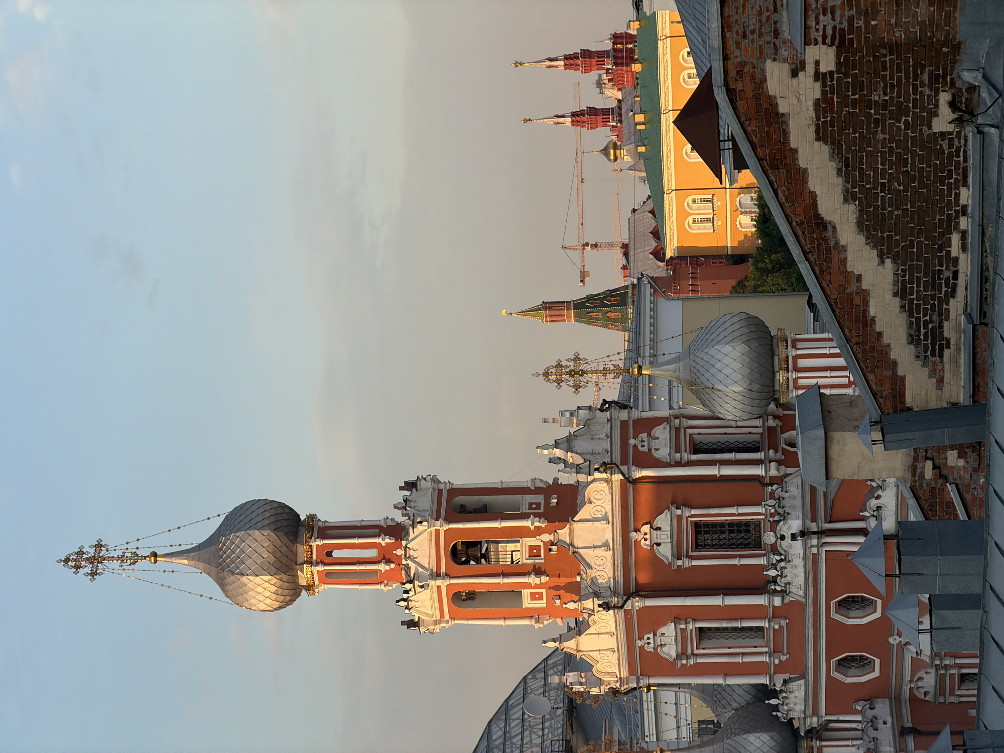 The image is a medium-shot, eye-level view of a picturesque scene in Moscow, Russia. In the foreground, a red and white church tower with a silver dome and a golden cross dominates the left side of the frame. The tower features intricate architectural details, including arched windows, decorative moldings, and bells visible inside. To the right of the tower, a rooftop with a brick wall and several metal chimneys is visible.
In the middle ground, a variety of colorful buildings with green and yellow roofs can be seen, interspersed with trees. Further in the background, several prominent Moscow landmarks are visible, including the iconic red towers of the Moscow Kremlin and a green-domed building, possibly a church. Construction cranes are also present, suggesting ongoing development or renovation in the city. The sky above is a soft blue with a few wispy clouds, bathed in the warm light of what appears to be late afternoon or early evening. The overall impression is one of historical grandeur and urban vibrancy.