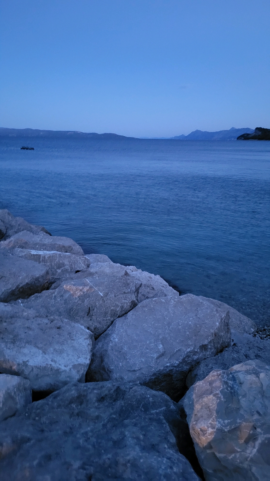 The image captures a twilight scene along the coast of Općina Tučepi, Croatia. In the foreground, large, rough, light-colored rocks form a breakwater or embankment, leading the eye towards the calm, dark blue sea. The water's surface shows subtle ripples, reflecting the fading light of the sky. In the distance, a chain of low-lying, dark hills or mountains stretches across the horizon, their forms softened by the low light. The sky above is a gradient of deep blue, transitioning to a lighter hue near the horizon. No people or specific structures are visible, emphasizing a natural, serene coastal landscape. The overall impression is one of a quiet evening at the edge of the Adriatic Sea.
