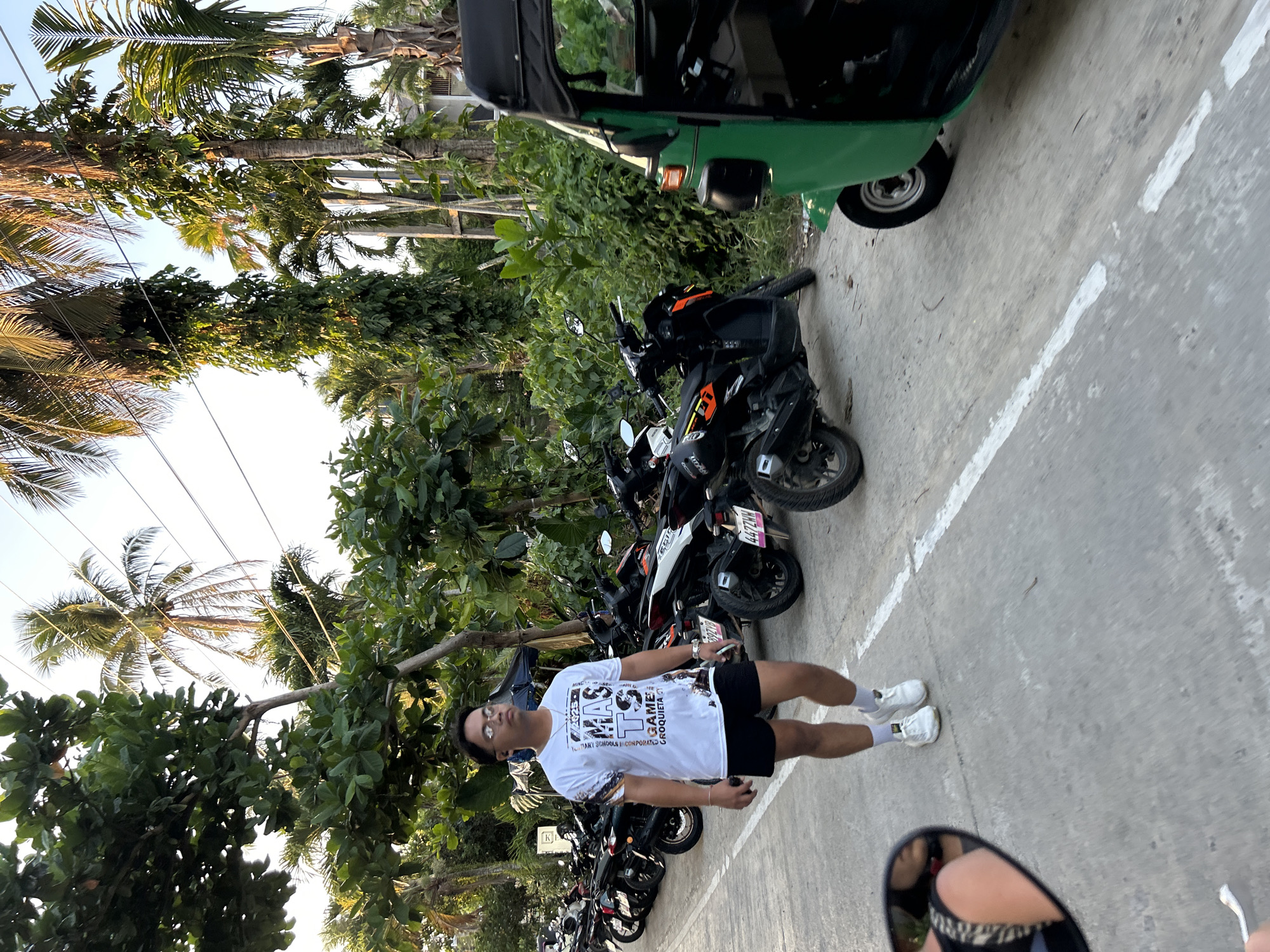 A daytime image shows a man standing on concrete near a row of parked motorcycles and a green motorized tricycle. The location is suggested to be in General Luna, Philippines. The man wears glasses, a white t-shirt, black shorts, white socks, and white shoes. He stands perpendicular to the camera's view. A row of motorcycles lines the left side of the image, and a green motorized tricycle is parked on the right side of the street. Tropical foliage and palm trees line the left side of the road. A partial view of another vehicle's mirror is in the bottom right corner. The ground is marked with white lane lines.
