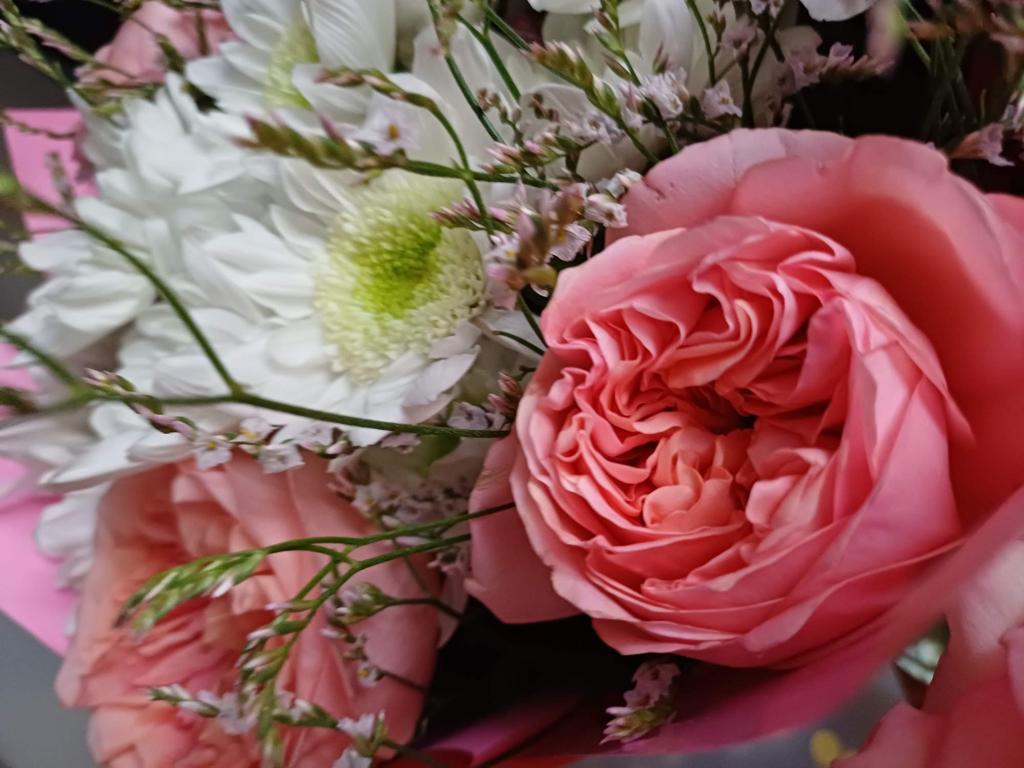 A close-up shot showcases a bouquet of flowers with predominantly pink and white hues. The foreground is dominated by a large, intricately layered pink rose, its petals unfurling in a spiral. Behind it, white flowers with a vibrant green center, resembling chrysanthemums, are visible. Delicate sprigs with tiny pink and white flowers are interspersed throughout the bouquet, adding texture and visual interest. The background consists of a blurred pink wrap, hinting at the bouquet's presentation. The lighting is soft, highlighting the delicate textures and soft colors of the flowers. No people or specific location details are discernible in this image.