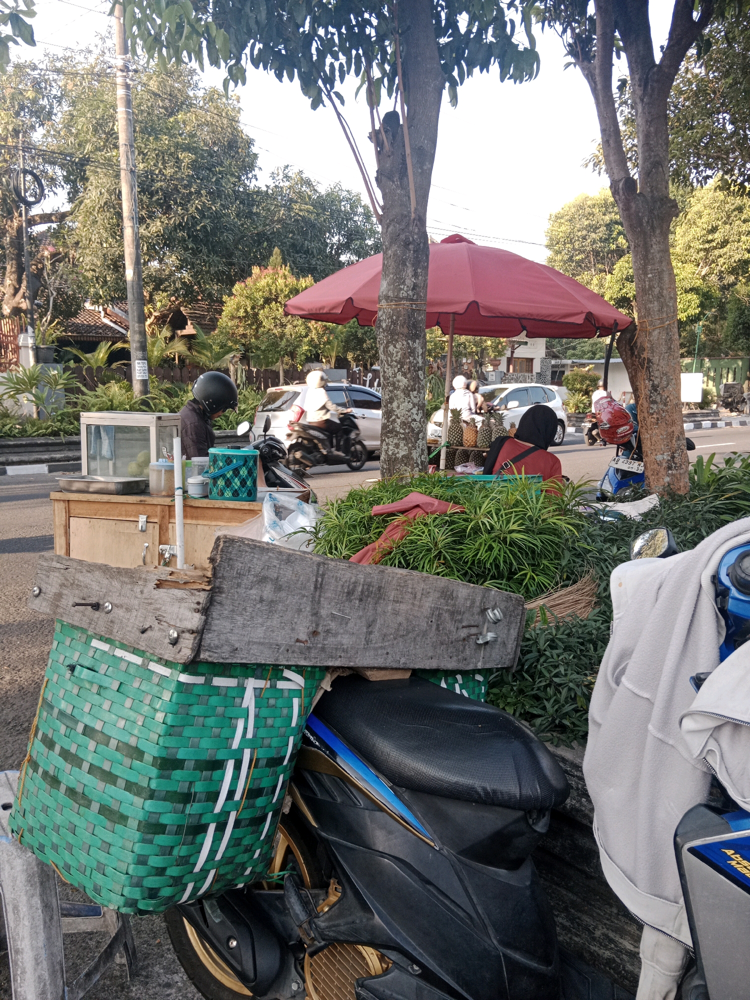 The image depicts a street scene in Yogyakarta, Indonesia, likely near a roadside market. In the foreground, a green woven basket is attached to a motorcycle. Next to the motorcycle is a wooden stall displaying a glass-enclosed food container and various items. A person wearing a black helmet is seen behind the stall, tending to the goods. Further back, a red umbrella shades a vendor selling pineapples and other items. Several motorcycles and a white car are visible on the road, indicating traffic flow. The environment is characterized by trees, a sidewalk, and residential buildings in the background, suggesting an urban or suburban setting. The lighting suggests it is daytime, possibly late afternoon due to the warm tones.