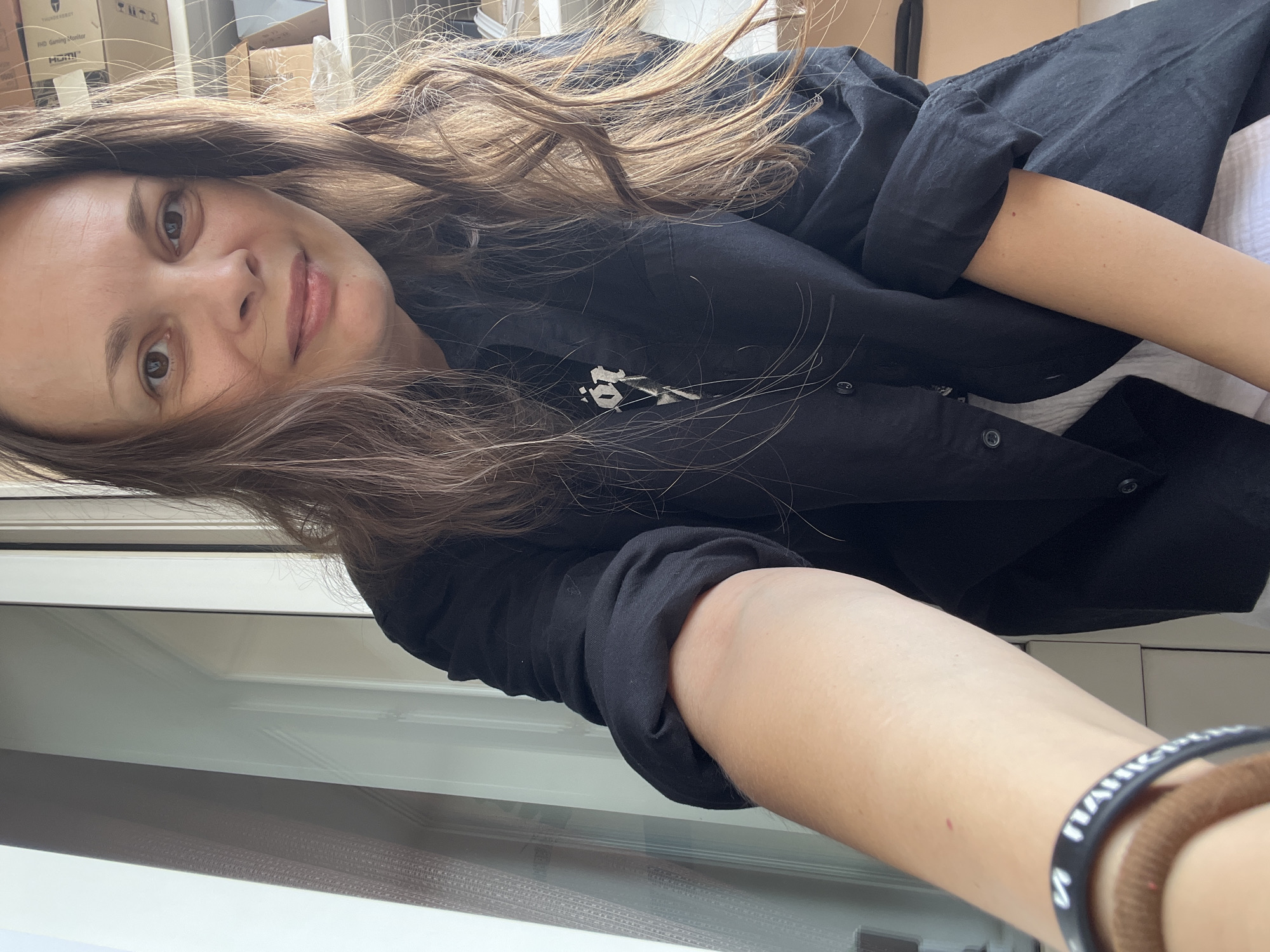 A woman is taking a selfie indoors, possibly on a balcony or near a large window. She has long, light brown hair and is wearing a black button-up shirt over a white garment. She is looking at the camera with a slight smile. The background shows a white door or window frame and some stacked boxes. The image is taken from a slightly upward angle, with her arm extended to capture the selfie. There is a black wristband with white writing on her left wrist, and a brown hair tie around her wrist as well. The scene suggests an informal setting. No specific location in Tolyatti, Russia, is identifiable from the image.