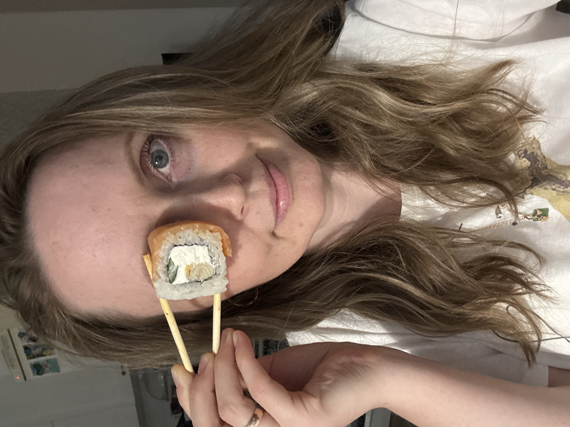 A woman with long, wavy blonde hair holds a sushi roll in chopsticks in front of her left eye. She is wearing a white t-shirt with a design on it. Her left eye is visible and appears to be light blue. She has a slight smile. She is wearing a wedding ring. The background is blurry and suggests an interior setting with some visible furniture. The photo may have been taken inside a house or apartment. There is no information in the image to indicate the location in the city of Chaikovsky, Russia.