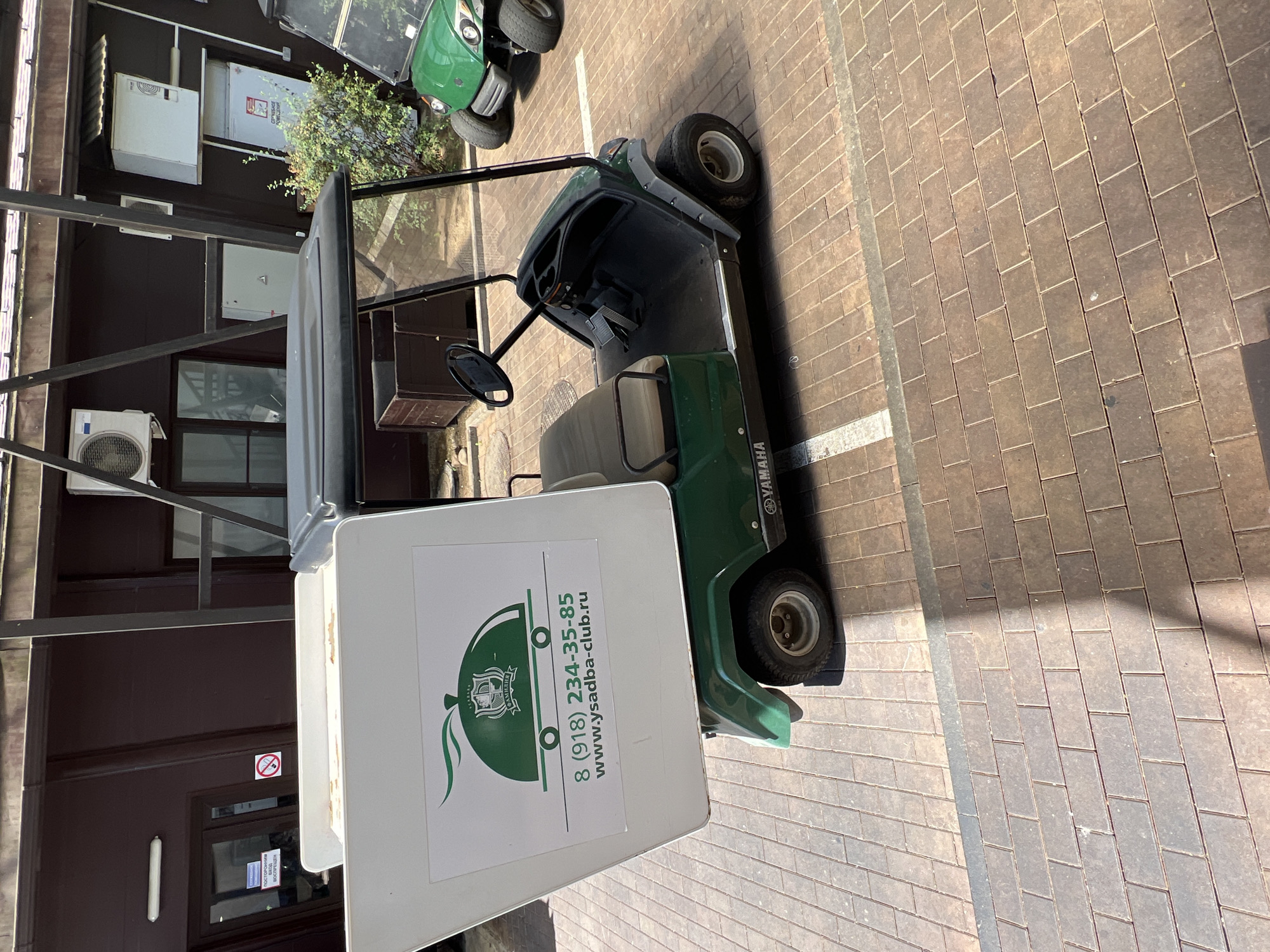 An outdoor, high-angle, eye-level shot shows a green Yamaha golf cart with a white cargo box attached to the rear, parked on a brick-paved area. The cargo box displays a logo with a cloche, a symbol of food service, and text that reads "8 (918) 234-35-85" and "www.ysadba-club.ru". The golf cart has a black roof and steering wheel, and a tan seat. A second, identical green golf cart is visible in the background, partially obscured by foliage. The scene appears to be in a commercial or recreational area with a building facade in the background, featuring dark brown siding and windows. The lighting suggests it is daytime, with some shadows cast on the ground.