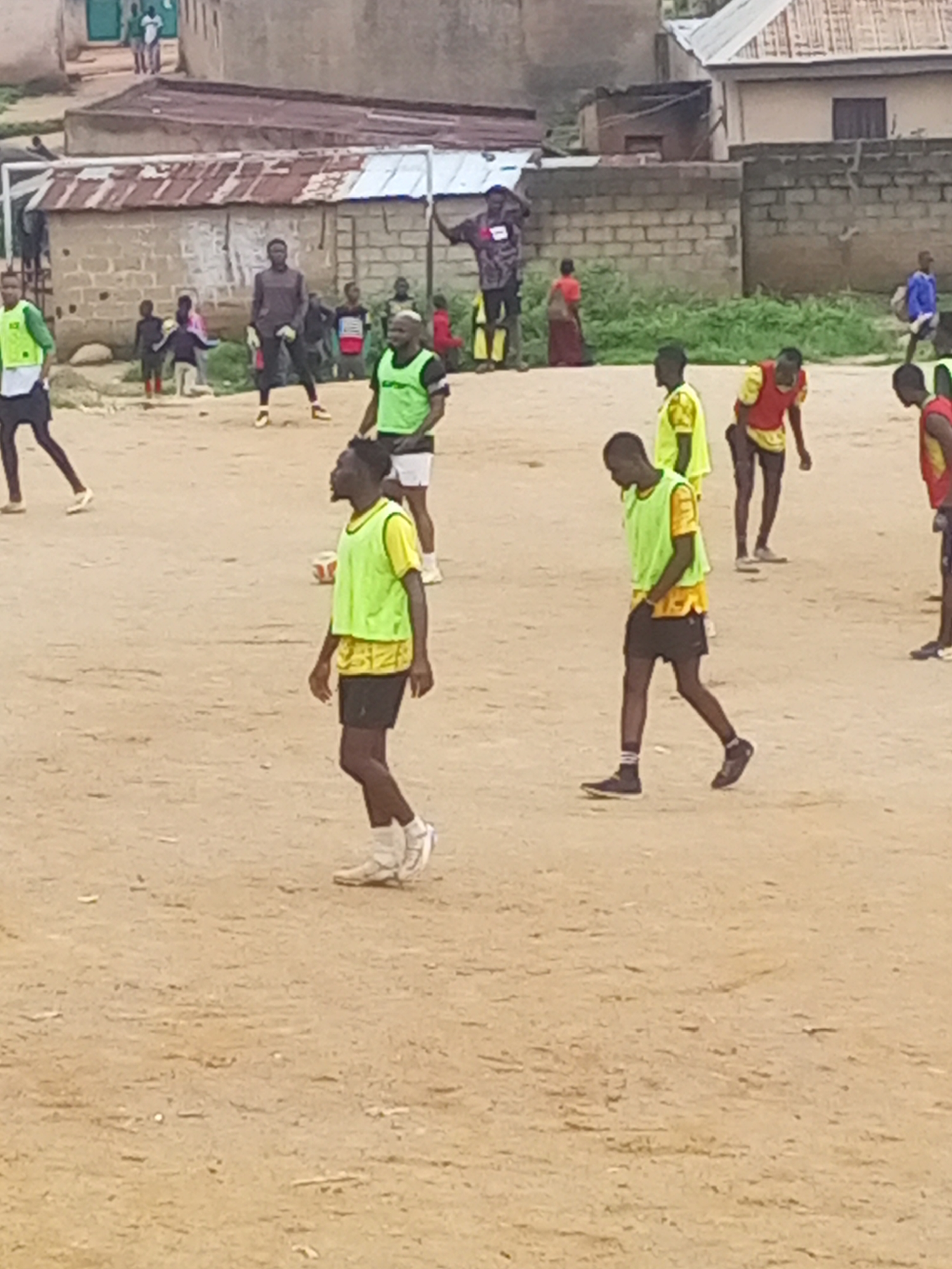 In Jos, Nigeria, a group of young men, likely a soccer team, are engaged in a practice session on a dusty, outdoor field. Most of the individuals are wearing brightly colored bibs, predominantly yellow and green, over their athletic attire. One person in the foreground, wearing a green bib, is looking to his left, with his hands down by his sides. In the background, other players are scattered across the field, some in motion and others observing. A soccer ball is visible on the ground near the center of the image. The environment suggests a casual, informal sporting event, with a backdrop of residential buildings and some vegetation. The overall impression is one of energetic youth engaged in physical activity in an urban setting.