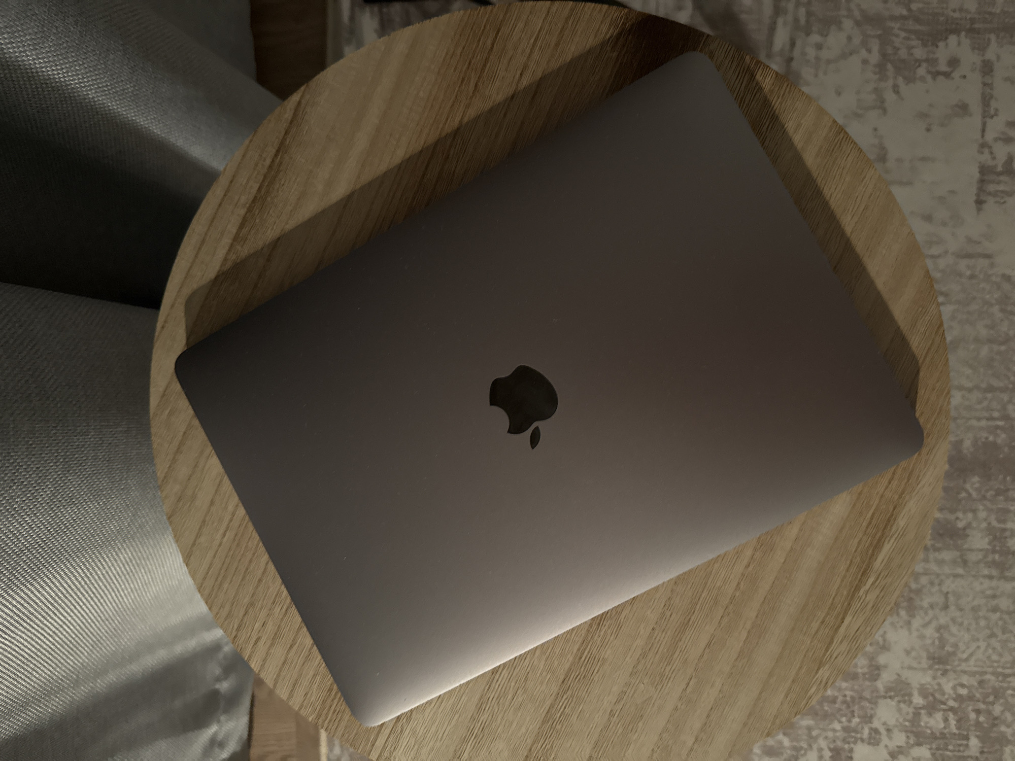 A dark gray Apple MacBook laptop is closed and lies on a round, light-colored wooden table. The Apple logo is visible in the center of the laptop lid. The table is placed on a textured rug with a pattern of gray and beige. In the background, a light gray, textured fabric, possibly a curtain or upholstery, is visible. The lighting suggests an indoor environment, potentially a home or office. The image does not provide any specific details indicating a location in Moscow, Russia.