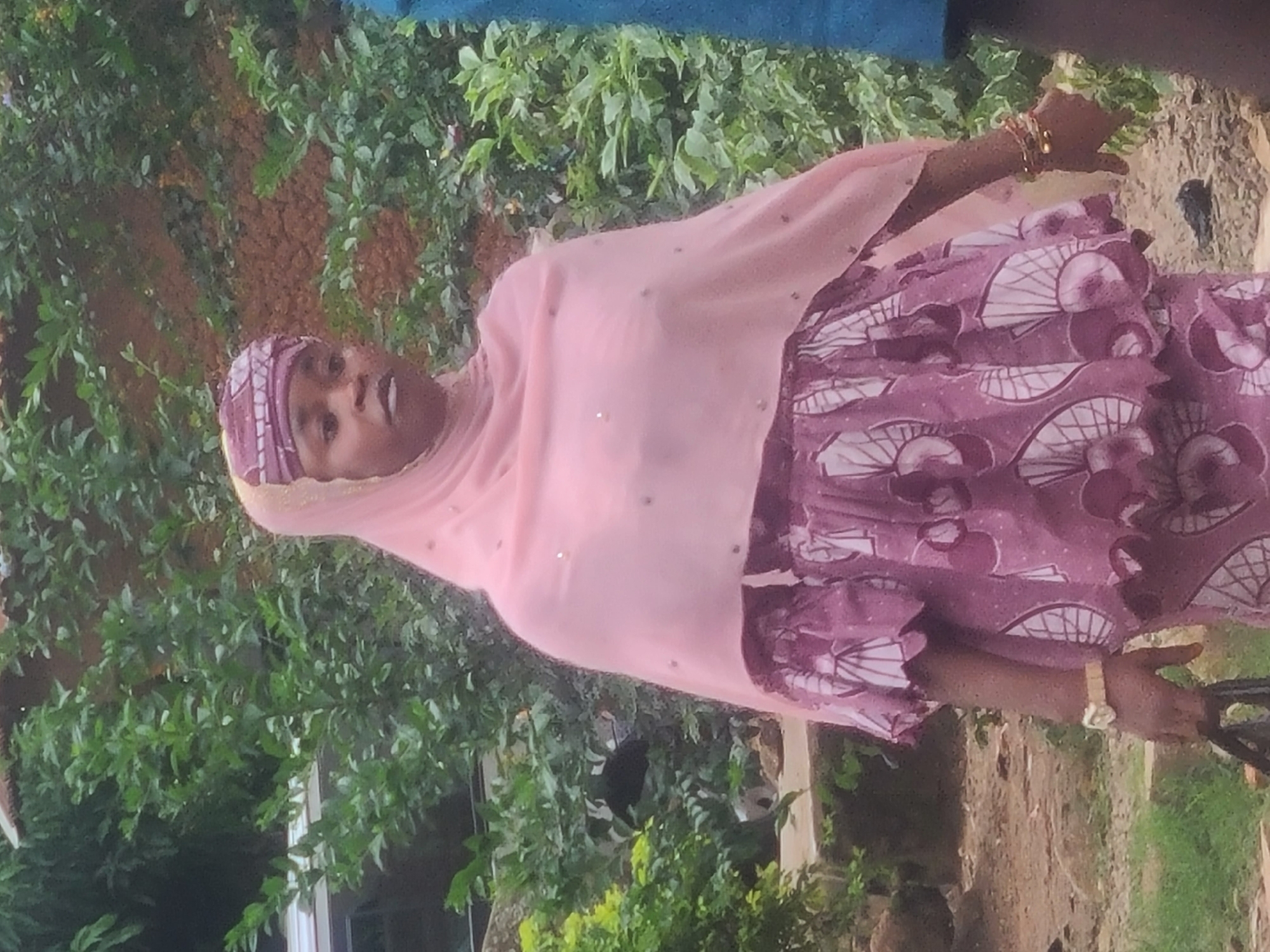 A woman, appearing to be of African descent, stands outdoors against a backdrop of lush green foliage and reddish-brown earth. She is wearing a pink headscarf with lighter pink accents and a matching pink shawl adorned with subtle embellishments. Her dress is a vibrant purple with a repeating white and pink circular pattern. A watch with a gold band is visible on her left wrist, and she has bracelets on her right wrist. She is holding a black bag in her left hand. The environment suggests a potentially rural or semi-urban setting within Jos, Nigeria, characterized by natural vegetation and a rough, unpaved ground surface. Another person, partially visible on the right side of the frame, is wearing a blue garment. The lighting indicates it is daytime, with soft, diffused light.
