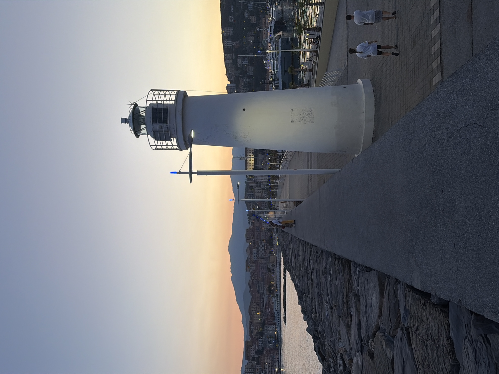 The image shows a lighthouse on a pier at dusk, with a coastal town and mountains in the background. Two people, likely teenagers, are walking away from the camera on the paved walkway. The lighthouse is white and cylindrical, with a metal cage around its lantern room. A single blue light is visible on a pole next to the lighthouse. The sky is a gradient of pale blue and orange, indicating sunset or sunrise. The town in the background is illuminated by lights, suggesting it is either late evening or early morning. The sea is calm and reflects the lights of the town. The pier is made of stone and concrete. The overall mood is peaceful and serene. The location appears to be Imperia, Italy, given the context.