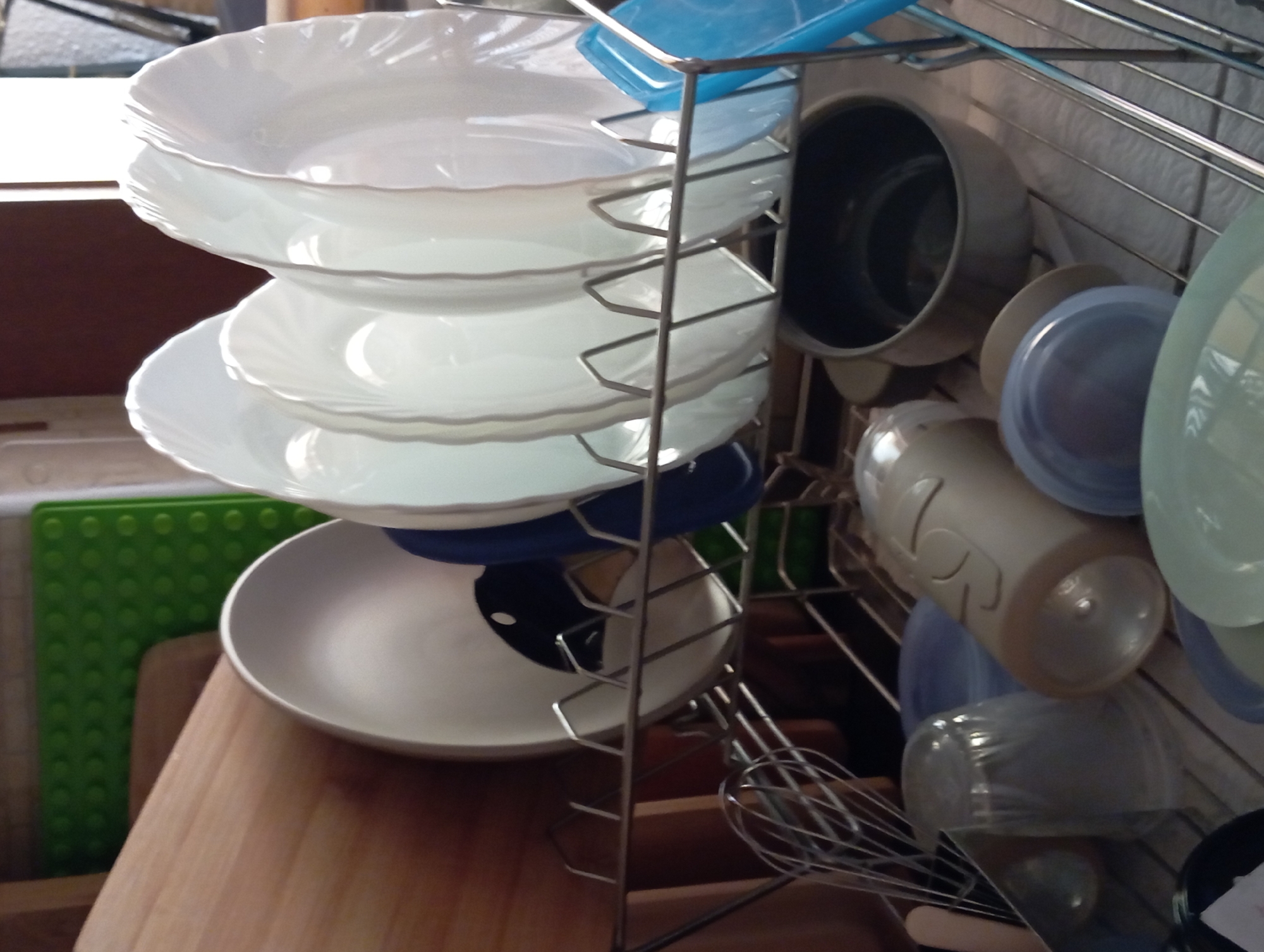 A close-up, eye-level shot shows a chrome dish rack filled with various white plates and bowls, some with fluted edges. The plates are stacked neatly, with the largest at the bottom. Beneath the plates, a blue bowl and a large, dark pot are visible. To the right, a beige plastic container with embossed markings, a blue bowl, and a lighter blue-green bowl are also present. Below these, a clear plastic container and a whisk are positioned. In the background, a green textured mat is partially visible. The setting appears to be a kitchen in Baden, Austria, suggesting the dish rack is either in use or being stored. The lighting indicates it is daytime.