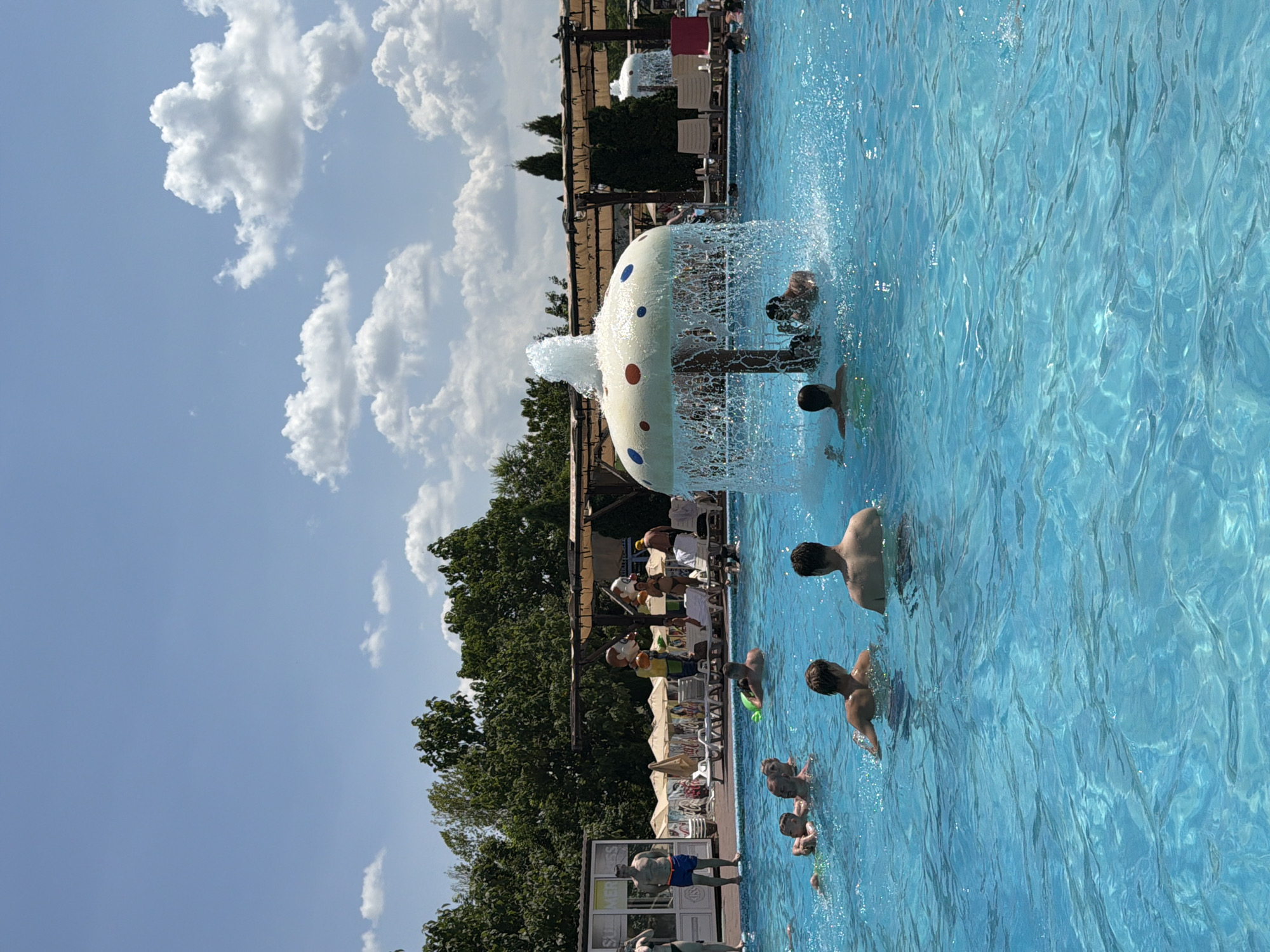 In the city of Sociteni, Moldova, a bright, sunny day is depicted at an outdoor swimming pool. The water is a vibrant blue, with ripples and splashes from the numerous people enjoying the refreshing water. In the center of the pool, a mushroom-shaped fountain sprays water, creating a playful and dynamic atmosphere. Several individuals are visible in the water, swimming, playing, and interacting. Some are partially submerged, while others are standing. In the background, trees and structures suggest a park or resort setting. The sky is a clear blue, dotted with fluffy white clouds, further enhancing the idyllic summer scene.