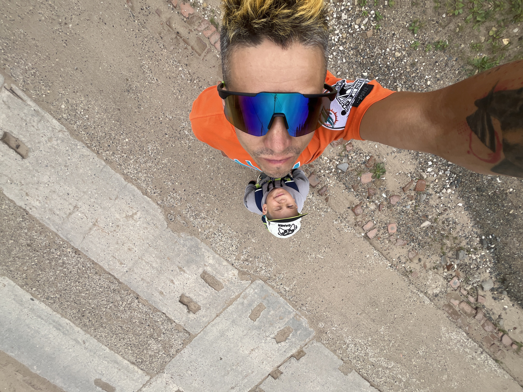 In Chaikovsky, Russia, a man and a young boy stand on a surface composed of concrete slabs and gravel. The man, positioned closer to the camera and looking upwards, wears an orange hooded sweatshirt with a Miami Dolphins logo and large, reflective sunglasses. His hair is spiked and dyed blonde at the tips. A tattoo is visible on his forearm. The boy, wearing a gray hooded shirt and a cap with "GAMER" printed on it, is positioned to the man's left and slightly behind him, also looking up. The ground beneath them is a mixture of textured concrete with embedded gravel and loose gravel mixed with broken bricks and dirt. The image is taken from a high angle, looking down at the subjects.