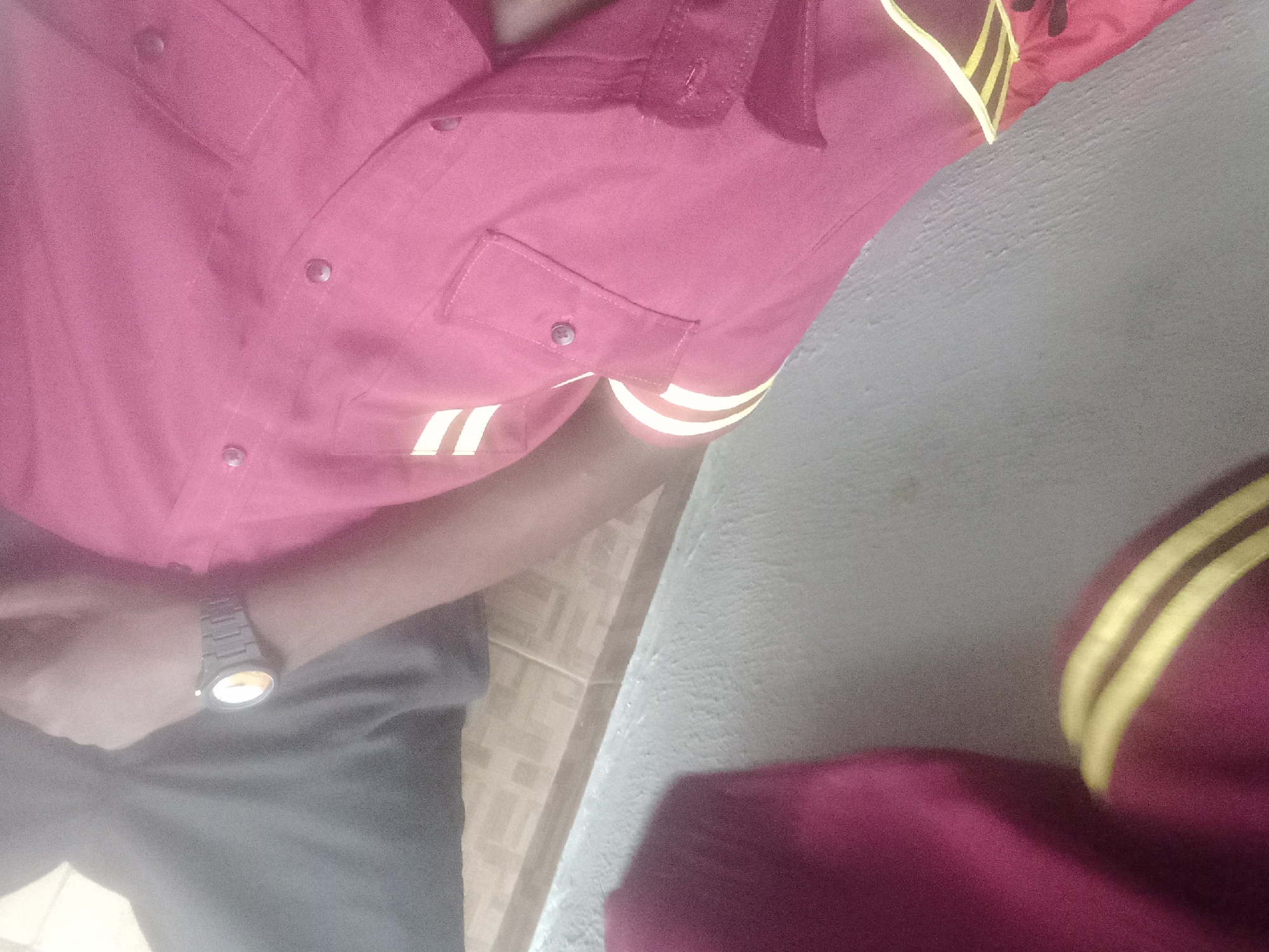 The image shows a close-up of a person wearing a maroon uniform with yellow stripes. The uniform appears to be a shirt with pockets and buttons. A watch is visible on the person's wrist. The background consists of a textured wall and tiled flooring. The scene is indoors, possibly in a residential or office setting. There is no explicit indication of the location being in Ilado, Nigeria, nor any specific actions or interactions depicted.