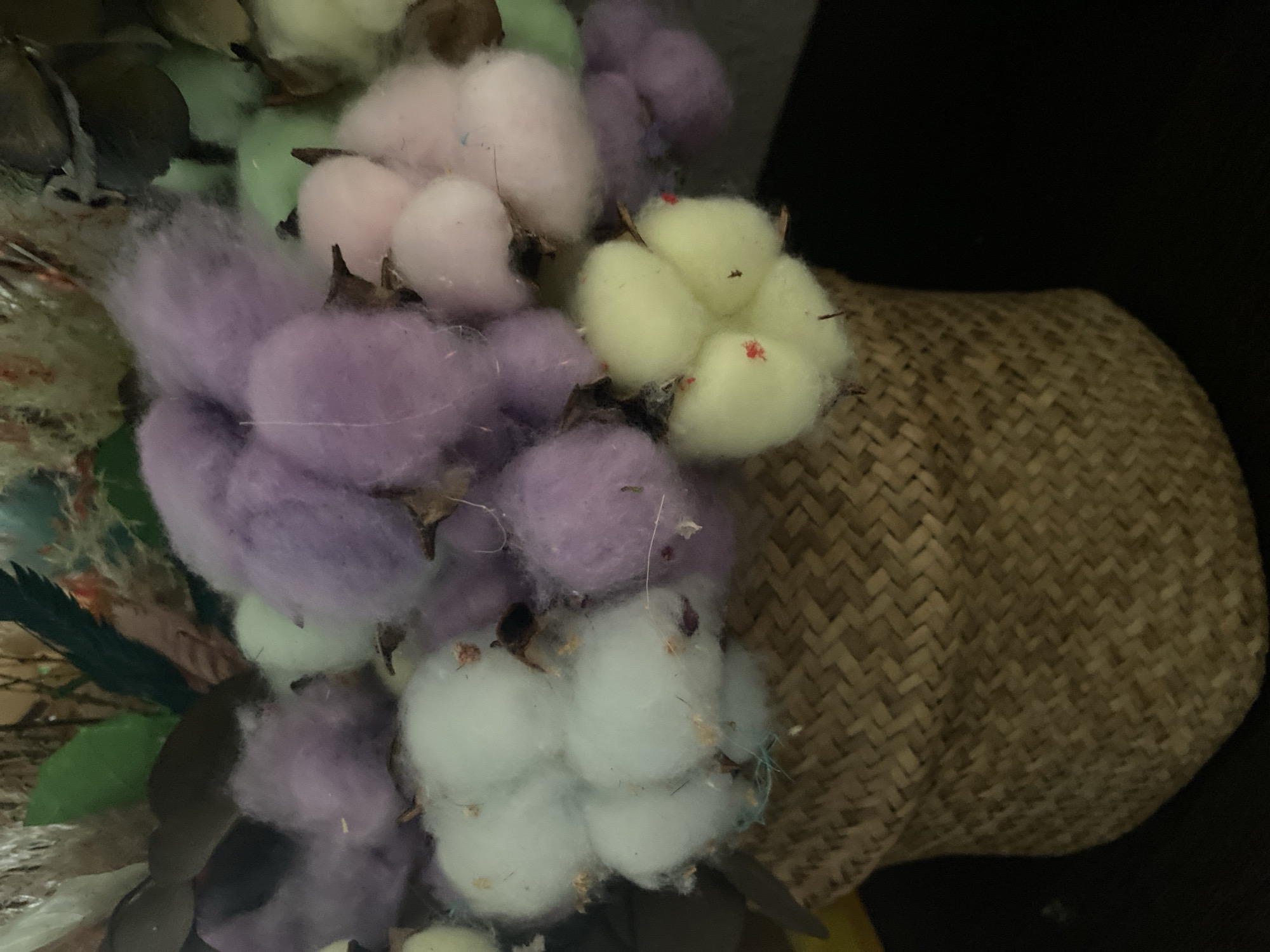 The image shows a close-up of a decorative arrangement featuring dyed cotton bolls in a woven basket. The cotton bolls are dyed in shades of lavender, pink, pale green, and yellow. The basket has a herringbone weave pattern and is positioned to the right of the cotton bolls. Some dried floral elements with green leaves are partially visible on the left side of the image. The background is dark and blurry. There is no evidence to suggest the location is Yekaterinburg, Russia. The image is devoid of people or actions.