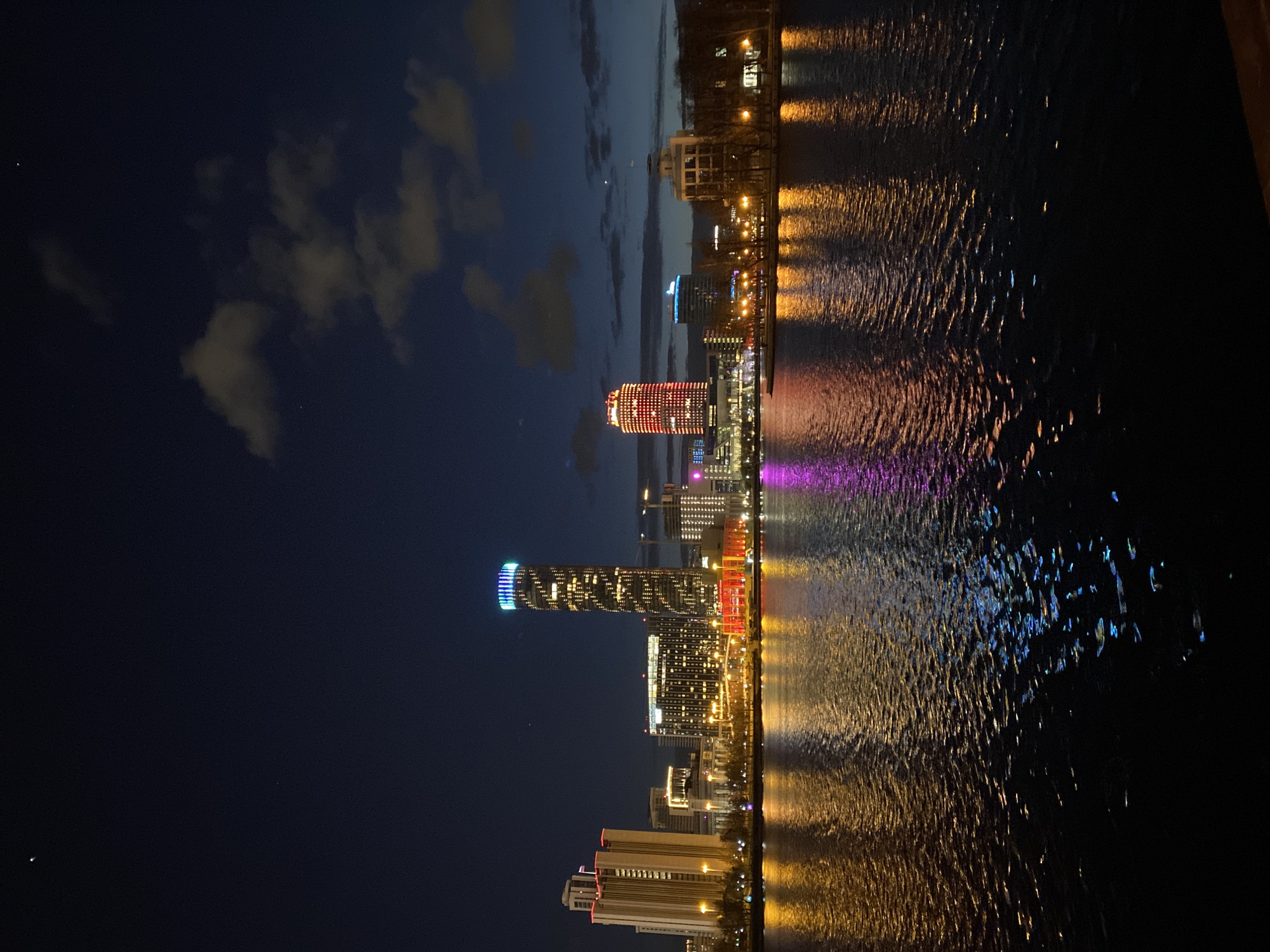 The image captures a nighttime view of the city of Yekaterinburg, Russia, reflected in a body of water. Prominent illuminated buildings line the waterfront, with their vibrant colors casting colorful reflections on the water's surface. The scene suggests an urban environment with modern architecture.