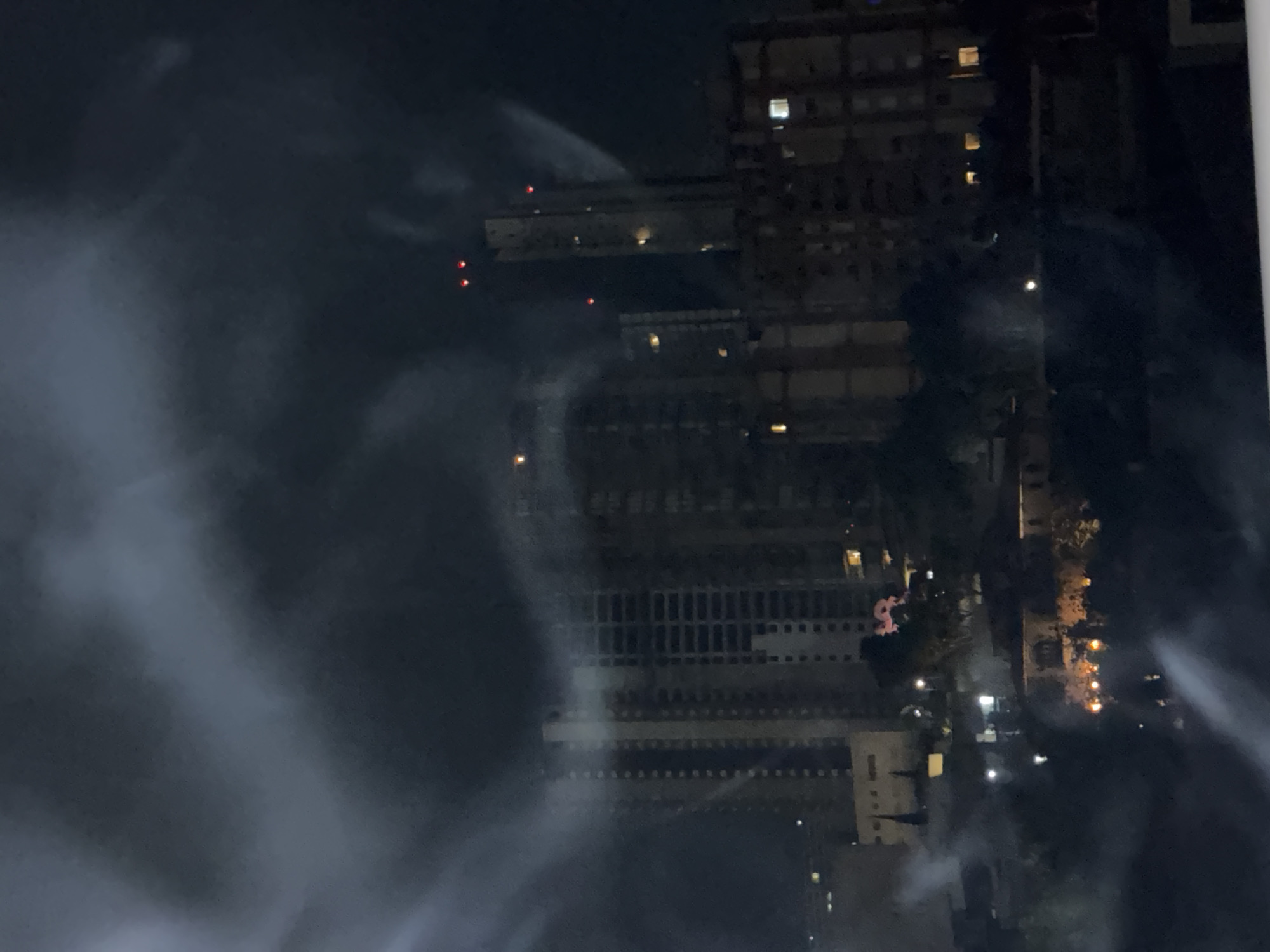 The image shows a nighttime view of buildings in Huixquilucan de Degollado, Mexico. High-rise buildings with lit windows dominate the scene. Heavy fog or smoke obscures parts of the view. Some lights appear to be streetlights or building lights. Red lights are visible on the top of some buildings.