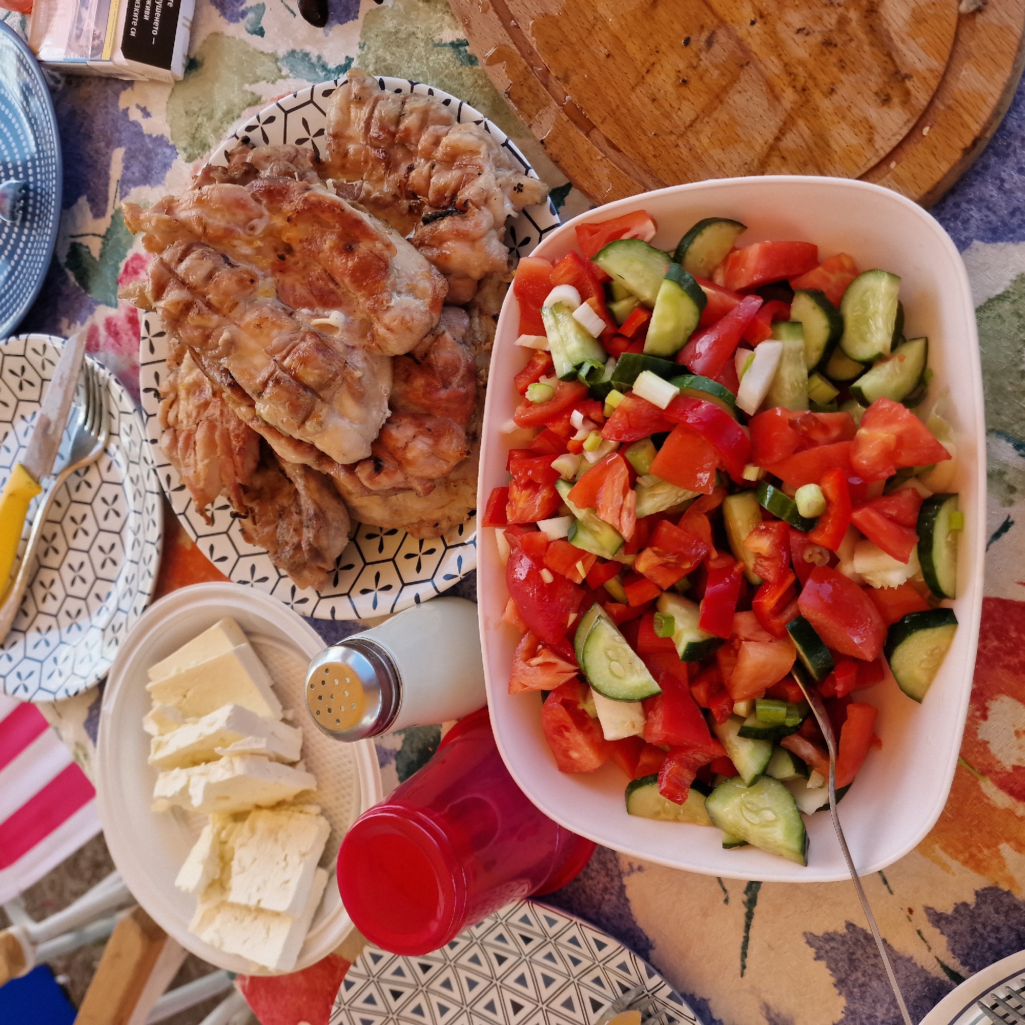 The image depicts a close-up view of a meal laid out on a table, possibly during an outdoor gathering in Gorni Dabnik, Bulgaria. In the foreground, a white bowl is filled with a fresh salad consisting of chopped tomatoes, cucumbers, red bell peppers, and green onions. A fork rests in the salad, suggesting it is ready to be eaten. To the left of the salad bowl, a red plastic cup is visible. Behind the salad and cup, on a plate with a geometric pattern, are several slices of white cheese. Further back, another plate displays grilled chicken. A salt shaker with a metallic top is positioned between the cheese and the grilled chicken. In the upper right corner, a pack of cigarettes is partially visible. The table is covered with a colorful tablecloth featuring floral and abstract patterns. The overall scene suggests a casual meal with a focus on fresh ingredients and grilled food.
