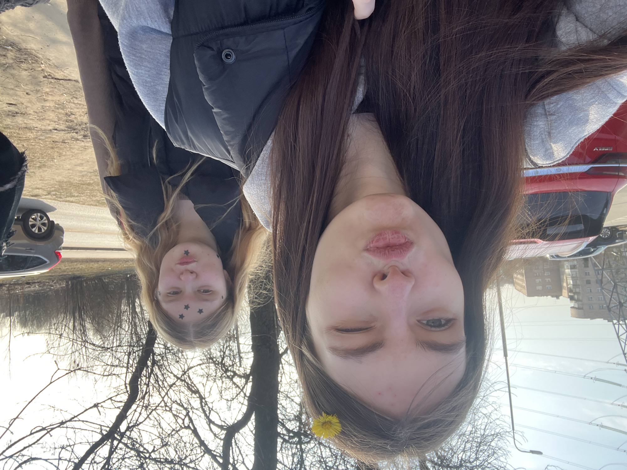 Two young women are posing for a selfie outdoors on a sunny day. One woman has long brown hair and a yellow flower tucked behind her ear. The other has long blonde hair and black star stickers on her forehead. Both are wearing jackets. In the background are bare trees, buildings, power lines, parked cars, and a road. The buildings suggest an urban environment, possibly in Saint Petersburg, Russia.