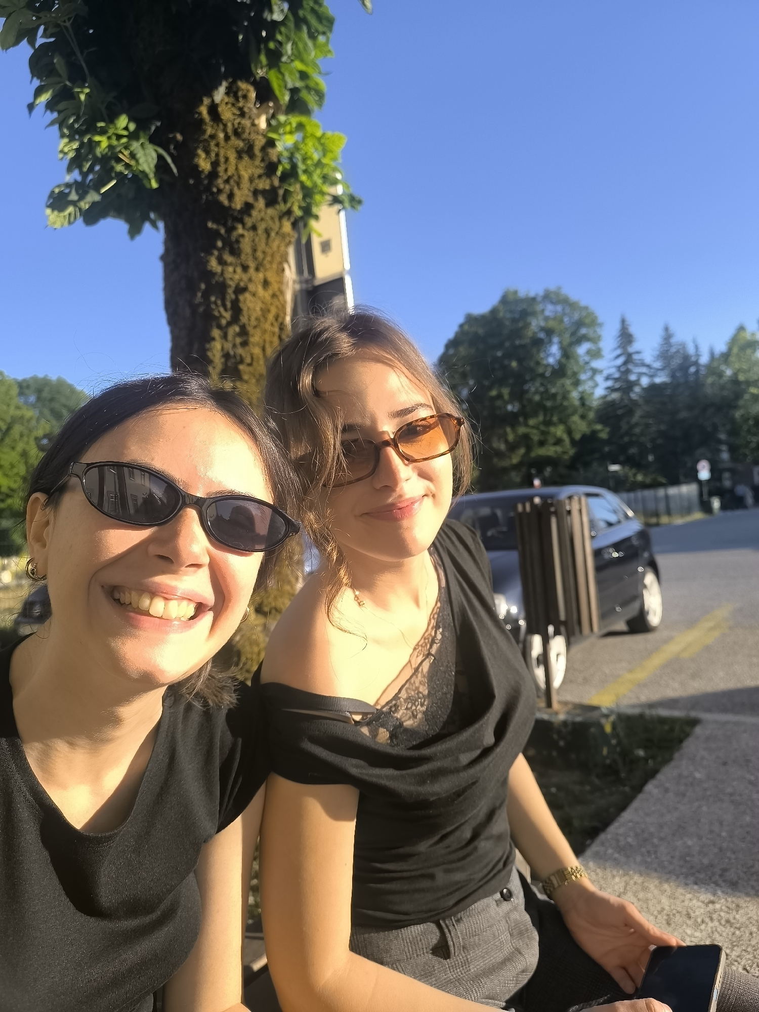 Two women are seated outdoors in a sunlit environment, with a tree to their left and a street with a car in the background. The woman on the left, wearing black sunglasses and a black t-shirt, smiles broadly, showing her teeth. The woman on the right, wearing orange-tinted sunglasses and a black off-the-shoulder top with lace detailing, also smiles. She is holding a smartphone in her right hand. The ground appears to be paved with concrete and there is a yellow line painted on the surface. The scene suggests a casual, possibly social, outing in an urban or suburban setting. The presence of a tree with moss on its trunk and the clear blue sky indicate a pleasant, possibly warm day. The architecture in the background is partially visible, suggesting buildings. The location is identified as the city of Cetinje, Montenegro.