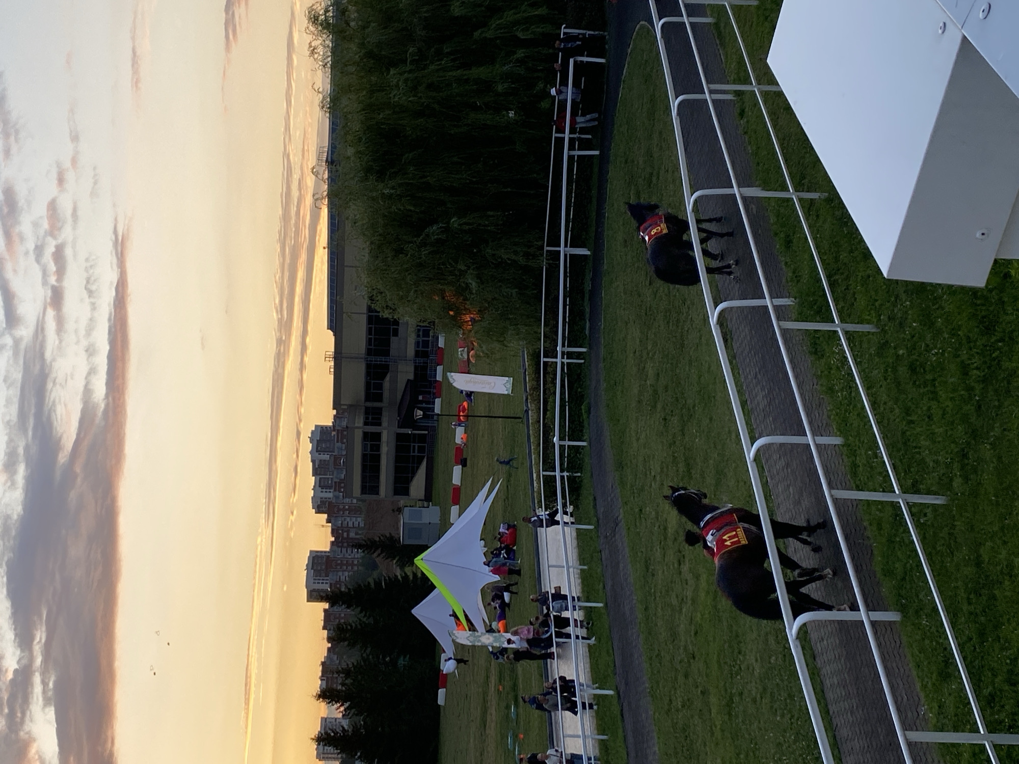 The image depicts a scene at a horse racing track at dusk, with the Kazan skyline visible in the background. The sky is filled with soft hues of orange, pink, and purple, indicating the sun has recently set. In the foreground, two horses wearing red racing blankets with numbers "11" and "8" are walking on a track. The track is bordered by a white railing and surrounded by green grass.
Further back, a grassy field contains several white, pyramid-shaped tents with green accents. People are gathered around the tents, some sitting on the grass and beanbag chairs. In the distance, there are trees and a large building. The overall atmosphere suggests an outdoor event or gathering taking place at a racecourse in Kazan, Russia.