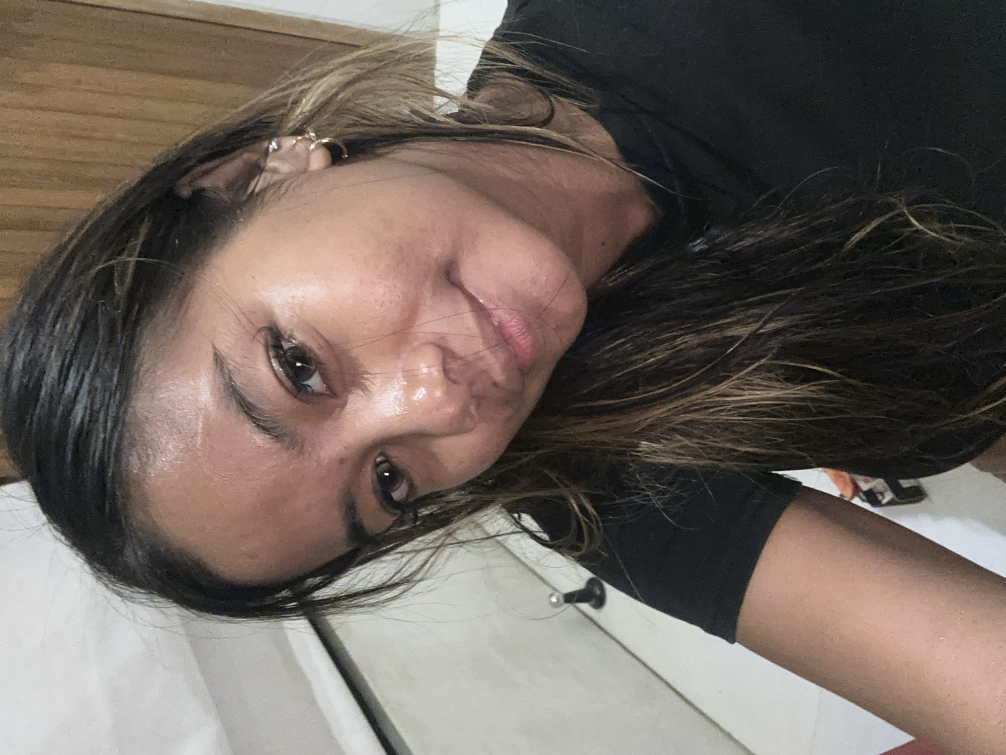A close-up, rotated image depicts a woman with dark hair streaked with lighter brown highlights. She is wearing a black shirt. She has visible ear piercings on her left ear. Her skin appears shiny, possibly due to sweat. The background includes a wooden door and a white wall. No information to indicate this is in Tumbak Bayuh, Indonesia.