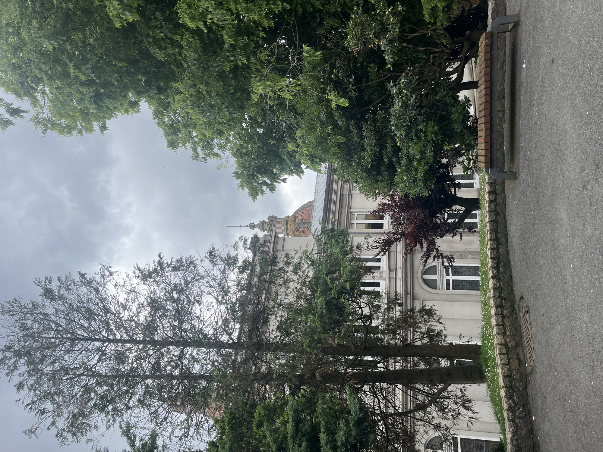 The image shows a building in A Coruña, Spain, partially obscured by trees. The building is multi-story and painted white, with several windows visible. It has a prominent dome on top, adorned with intricate patterns. The sky is overcast, and the street is lined with trees and a stone curb. The image appears to be taken from a low angle, capturing the building through the trees. The location is likely in a residential or institutional area due to the architecture and street setting.