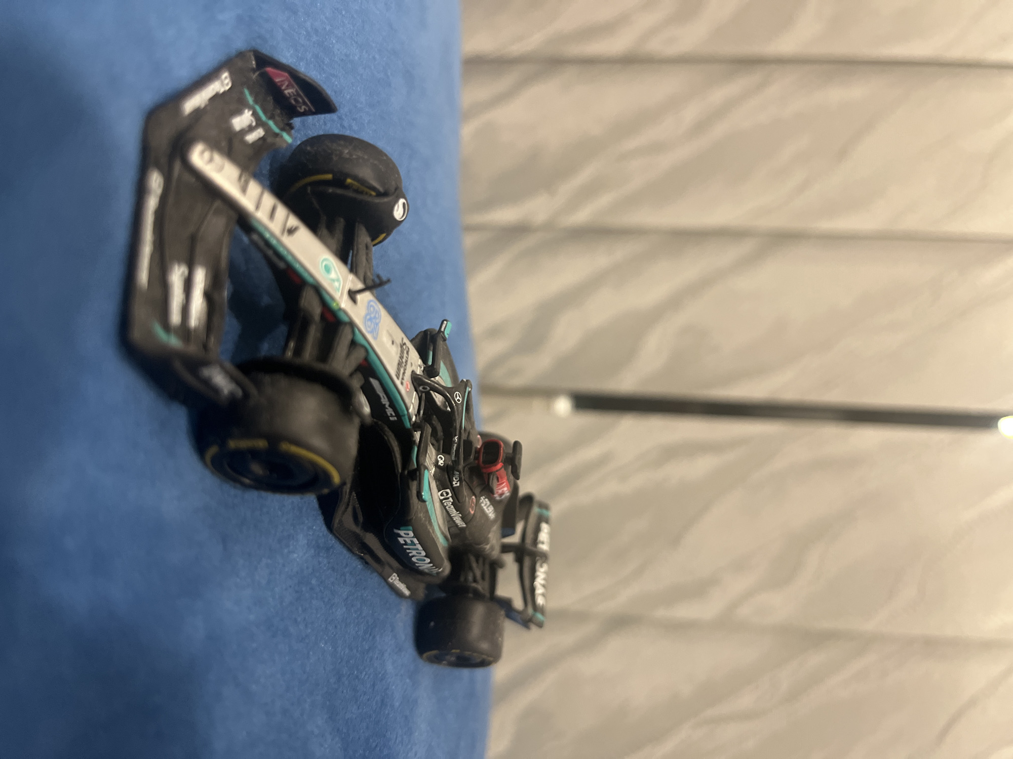 A toy model of a Mercedes Formula One car is lying on a blue surface. The car is black and silver with green accents, and various sponsor logos such as "Petronas", "TeamViewer", and "INEOS" are visible on the body. The background is a beige surface with horizontal lines. The image does not show any indication of Vladaya, Bulgaria.