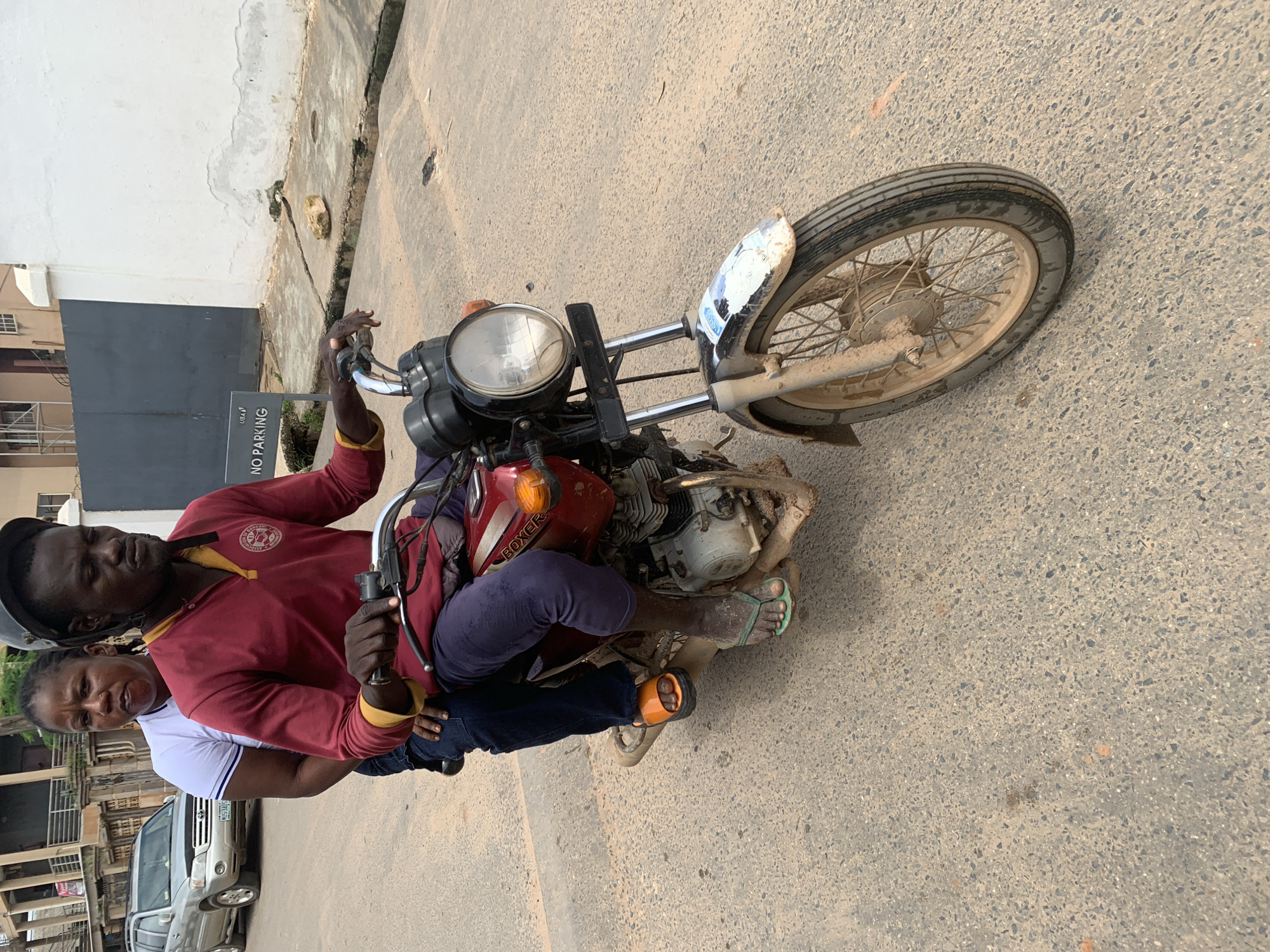 A man wearing a helmet and a maroon shirt is driving a red motorcycle with a woman wearing a white shirt as a passenger. They are on a paved road in Shagamu, Nigeria. The man is looking forward, and the woman is looking towards the camera. The motorcycle has a license plate, a headlight, and a passenger seat. The background shows buildings and a sign that says "NO PARKING". The road is dirty, with some debris scattered around.