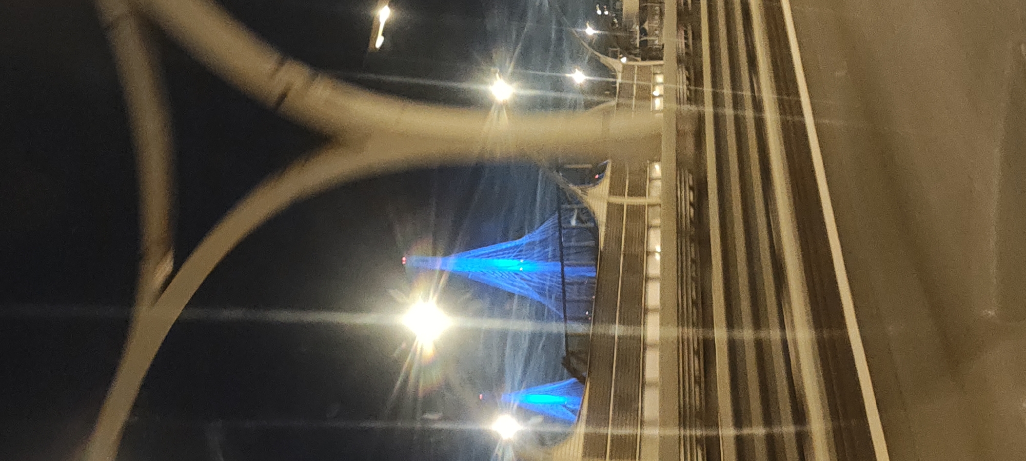 A vertical, medium shot captures the nighttime view of two illuminated cable-stayed bridges in Saint Petersburg, Russia, from the perspective of being on a road or elevated walkway. The dominant feature is the vibrant blue illumination of the bridge towers, which are shaped like inverted cones and are supported by numerous cables. The sky is dark, indicating it is nighttime, with some wispy clouds visible. In the foreground, a blurred concrete structure, likely part of a road or bridge infrastructure, frames the view. There are horizontal lines of light suggesting railings or barriers, and below that, the blurred tracks of a road or railway. Several bright light sources, some with lens flare effects, are scattered across the scene, illuminating the foreground and parts of the bridge structures. The image appears to be taken from a moving vehicle due to the motion blur.