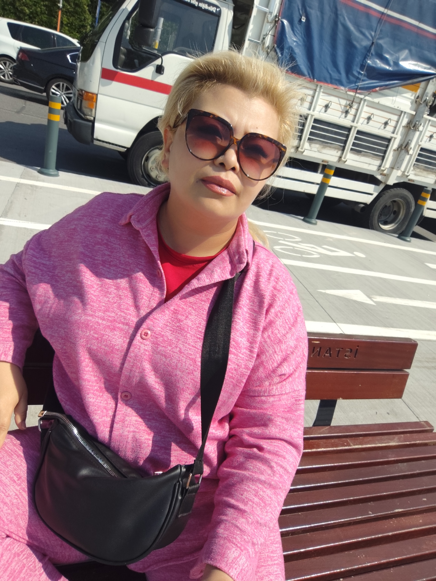 The image shows a blonde-haired woman wearing a pink outfit and sunglasses, sitting on a wooden bench. She is wearing a black crossbody bag. Behind her is a white truck with a blue tarp. The ground is gray with white arrows indicating a bike path. The bench has text on it in a foreign language, likely Turkish, and includes the word "İSTANBUL". This suggests the location is in Istanbul, Turkey. Considering it is Sarıyer, the bench is likely located in the Sarıyer district. There is a white car and smaller white truck further in the background.
