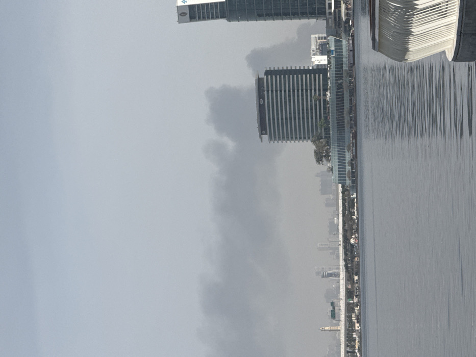 A panoramic view across Dubai Creek in Dubai, United Arab Emirates, depicts a significant environmental event. A large, dense plume of dark grey smoke extends horizontally across the mid-section of the sky, originating from an area beyond the visible structures on the right.
In the foreground, a body of rippled grey water occupies the lower half of the image. A white, curved, ribbed architectural element, likely part of a promenade railing or seating, is partially visible in the extreme bottom right corner.
The midground features the skyline of Dubai Festival City. On the far right, the InterContinental Dubai Festival City hotel is prominent, identifiable by its modern, multi-story structure and a logo near its apex. To its immediate left stands another multi-story building characterized by horizontal bands of windows, positioned above a lower, wider building with a glass facade and scattered rooftop palm trees. Further left across the creek, a bridge spans the water, with a slender, minaret-like structure visible near its base. Distant city buildings form the background skyline beyond the bridge.
The sky above the smoke plume is light grey, indicating either overcast conditions or atmospheric haze. No individuals or specific actions are discernible within the frame.