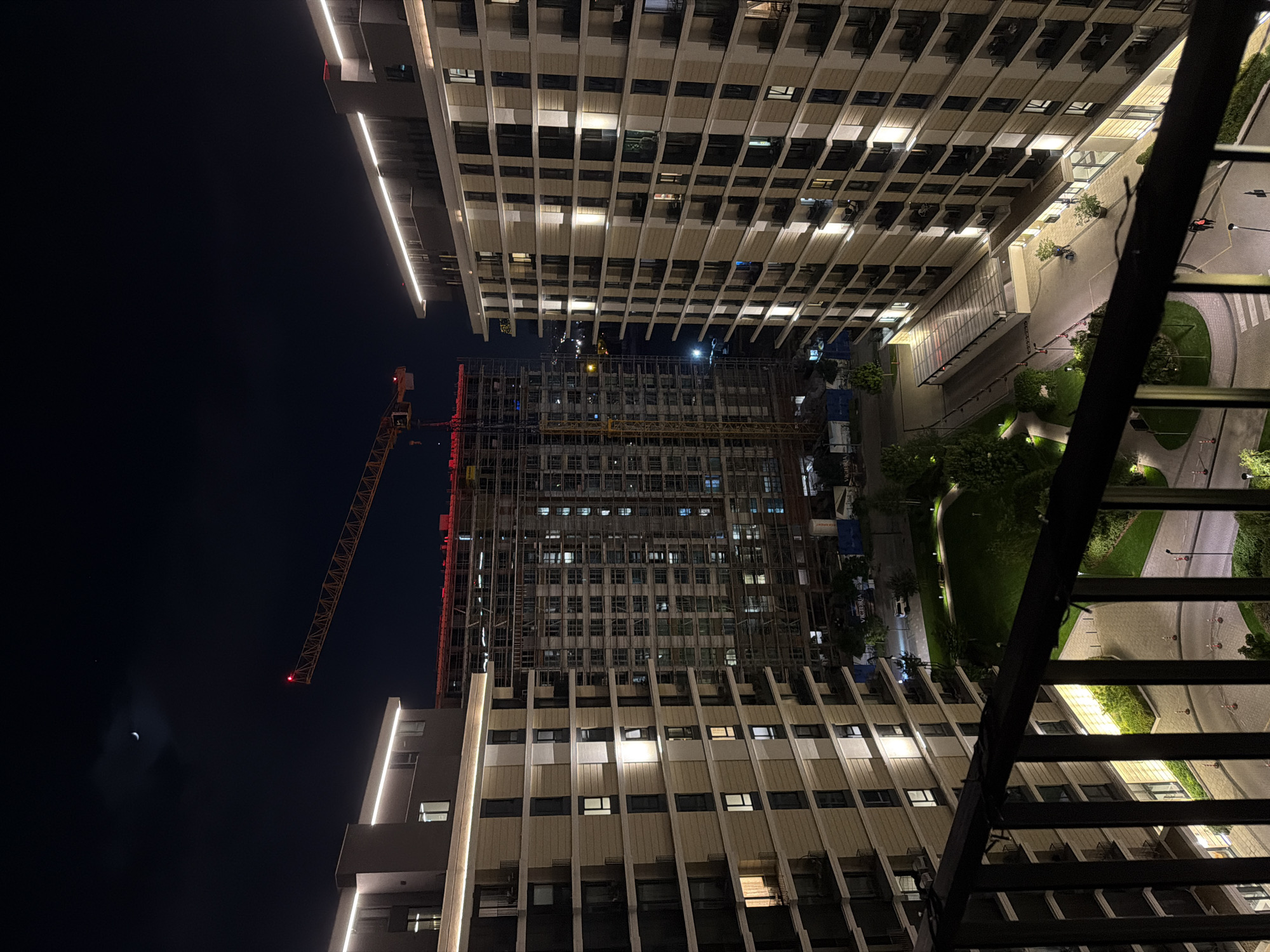 The image depicts an urban scene at night, likely taken from a high vantage point such as a balcony. Two tall residential buildings with lit windows flank a construction site where a new building is being erected. A yellow crane stands adjacent to the incomplete building. A park with paths and vegetation is visible on the right side of the frame. The moon is visible in the upper left corner. The location is Belgrade, Serbia.