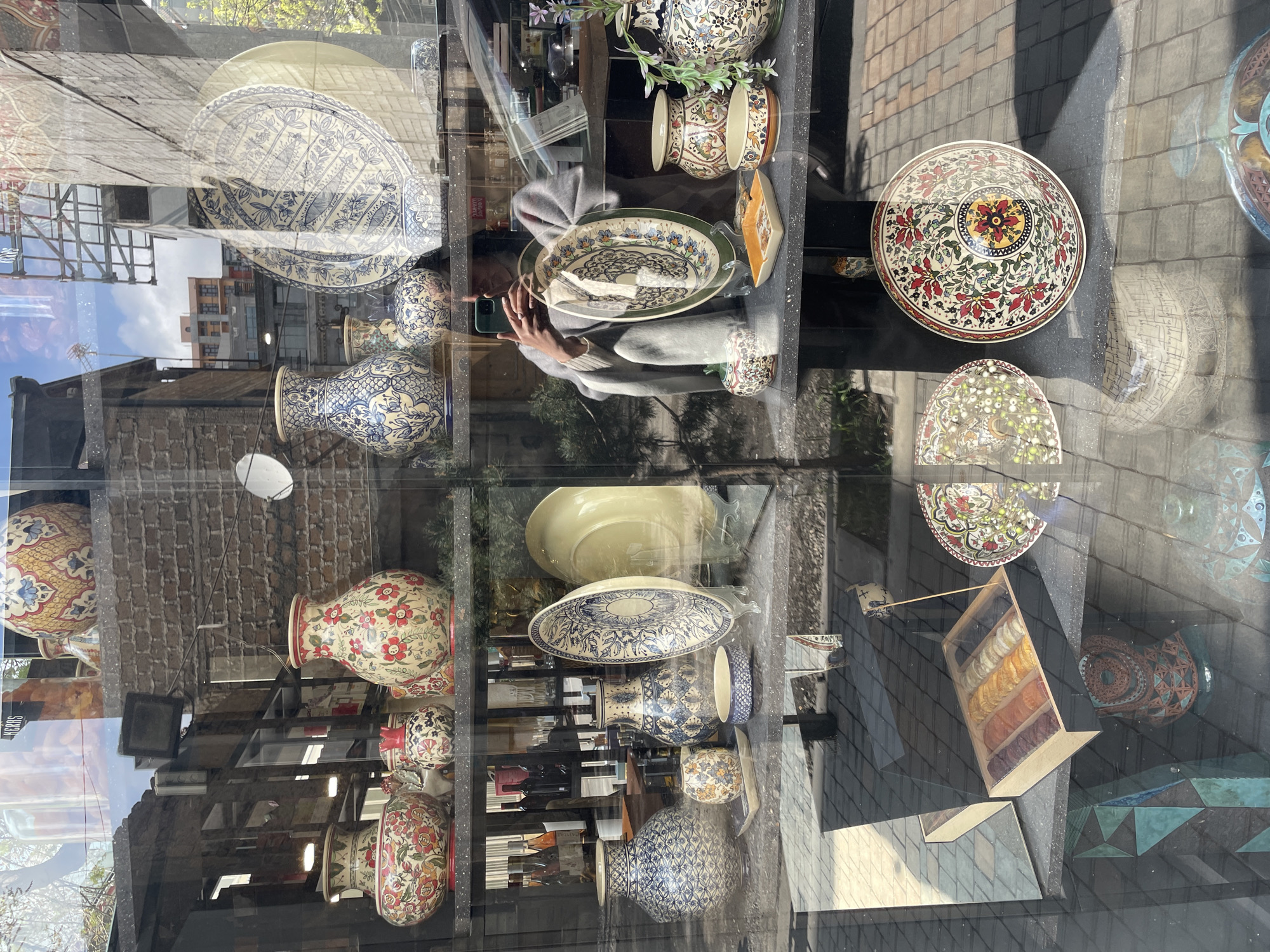 The image depicts a shop window in Yerevan, Armenia, displaying an array of artisanal pottery and ceramics. Visible in the window are vases, plates, and bowls, each decorated with intricate patterns in various color schemes, including floral motifs and geometric designs. The shelf arrangement is multi-tiered, showcasing the variety of items for sale.
The reflective glass of the shop window displays a street scene, including buildings under construction with scaffolding, other commercial establishments, and pedestrians. A person is reflected taking a photo of the display with a smartphone. The foreground shows a street scene with pavement.