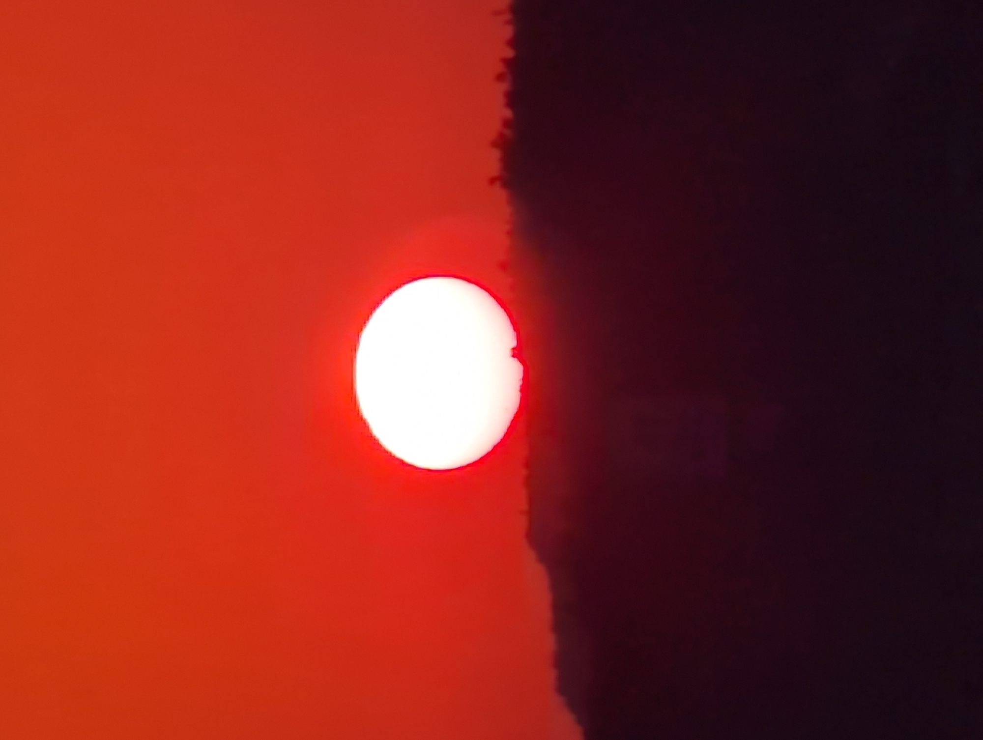The image depicts a sunset in Kryvyi Rih, Ukraine. The sun, a bright white disc with a reddish halo, is positioned to the left of the frame, partially obscured by a dark, textured silhouette on the right. This silhouette appears to be a tree line or the edge of a structure, casting a deep shadow across the right half of the image. The left side of the frame is dominated by a vibrant, warm orange glow, characteristic of a sunset, which gradually fades into a darker hue towards the bottom. The contrast between the brightly lit sun and the encroaching darkness suggests a dramatic and perhaps serene moment as day transitions into night. No people or specific objects are clearly discernible, the focus being on the natural phenomenon of the setting sun and its atmospheric effect on the landscape. The image captures a peaceful yet striking natural display, with the time of day being clearly evening. There is no visible text in the image.